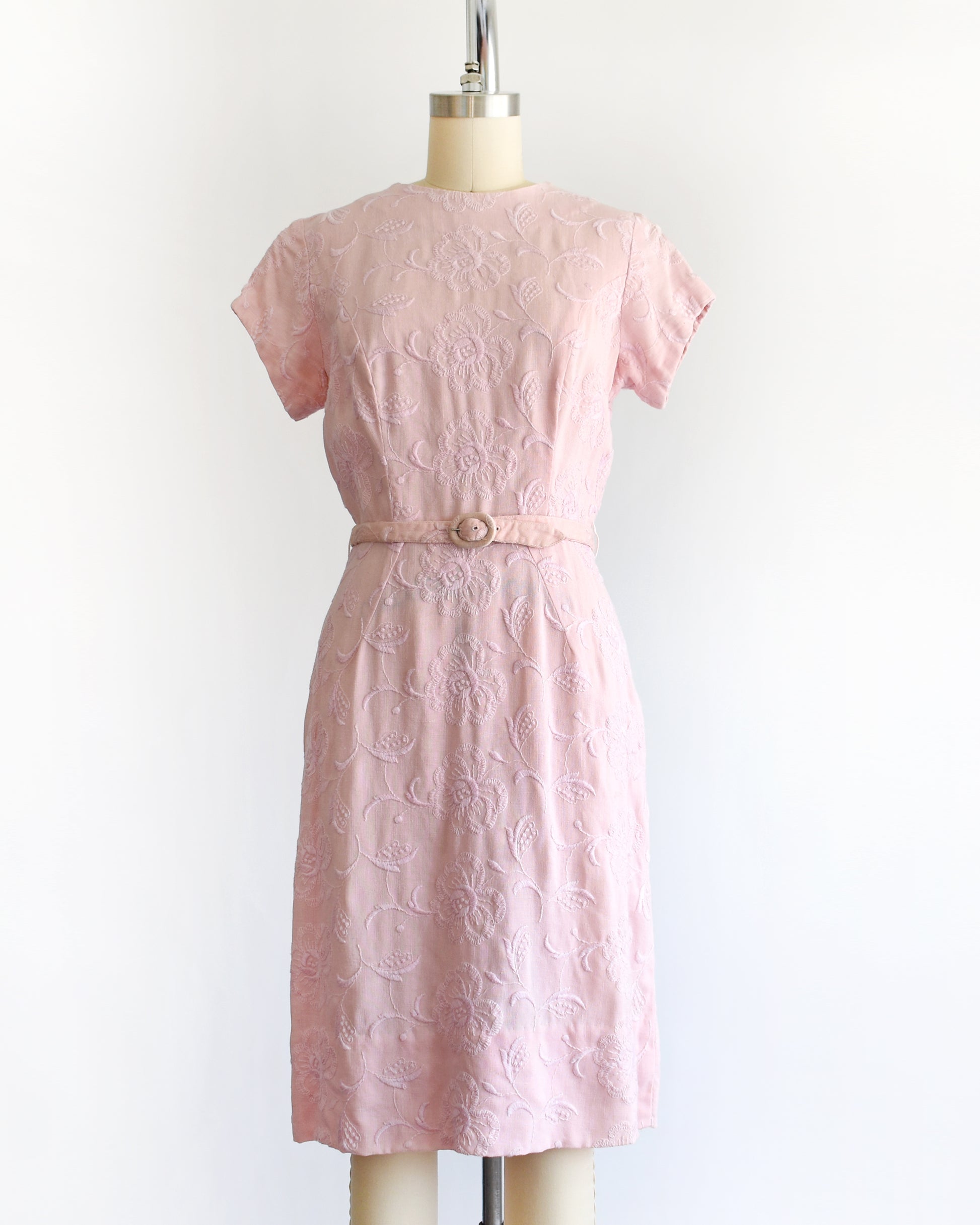 A vintage 1950s/1960s light pink short-sleeve dress with an embroidered floral pattern and a matching waist belt, displayed on a cream-colored dress form against a white background.