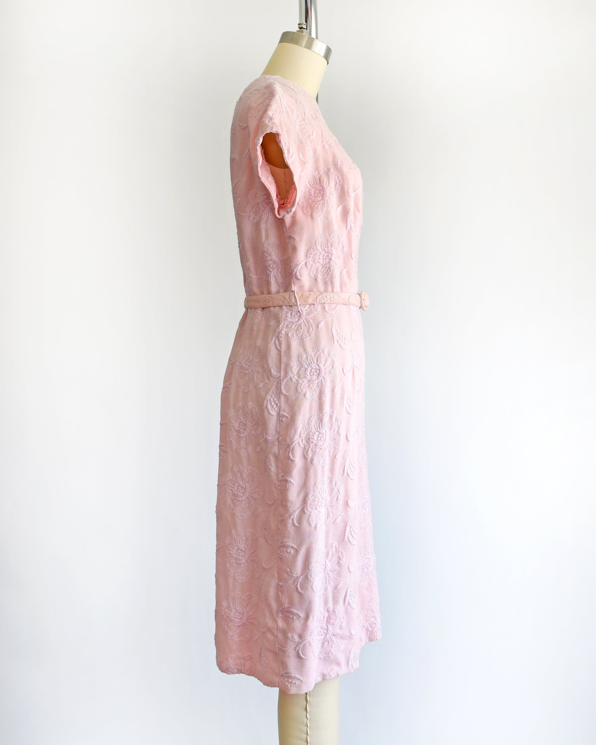 side view of a vintage 1950s/1960s light pink short-sleeve dress with an embroidered floral pattern and a matching waist belt, displayed on a cream-colored dress form against a white background.