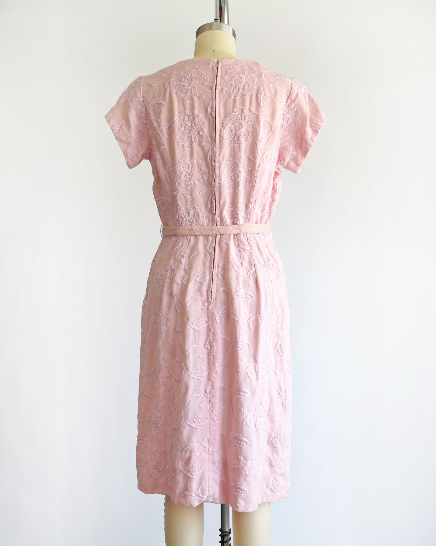 back view of a vintage 1950s/1960s light pink short-sleeve dress with an embroidered floral pattern and a matching waist belt, displayed on a cream-colored dress form against a white background.