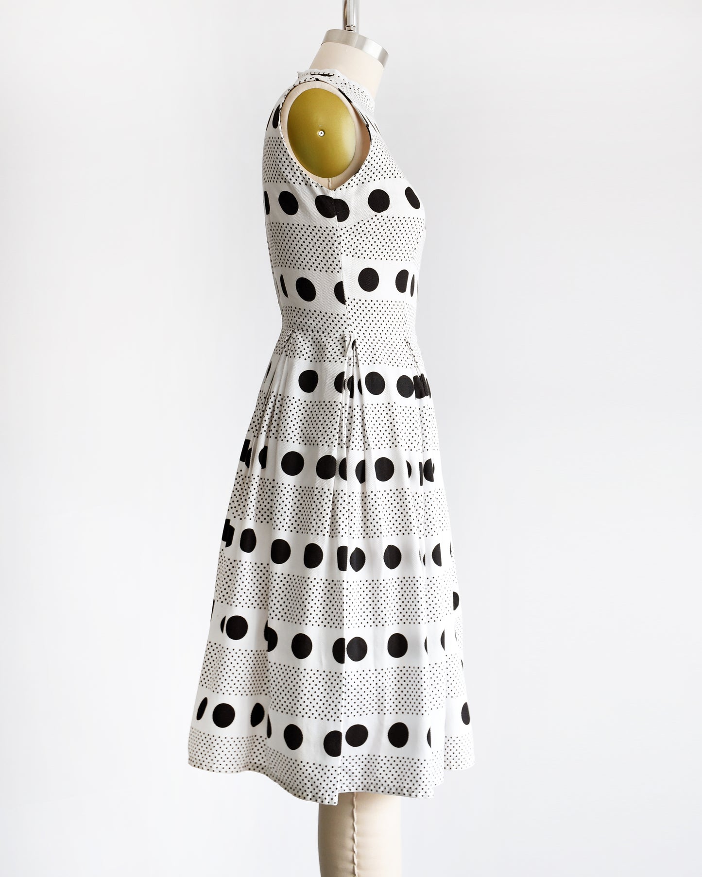 side view of a vintage 1960s polka dot dress that is white cotton piqué with large and small black polka dots in a striped pattern on a dress form