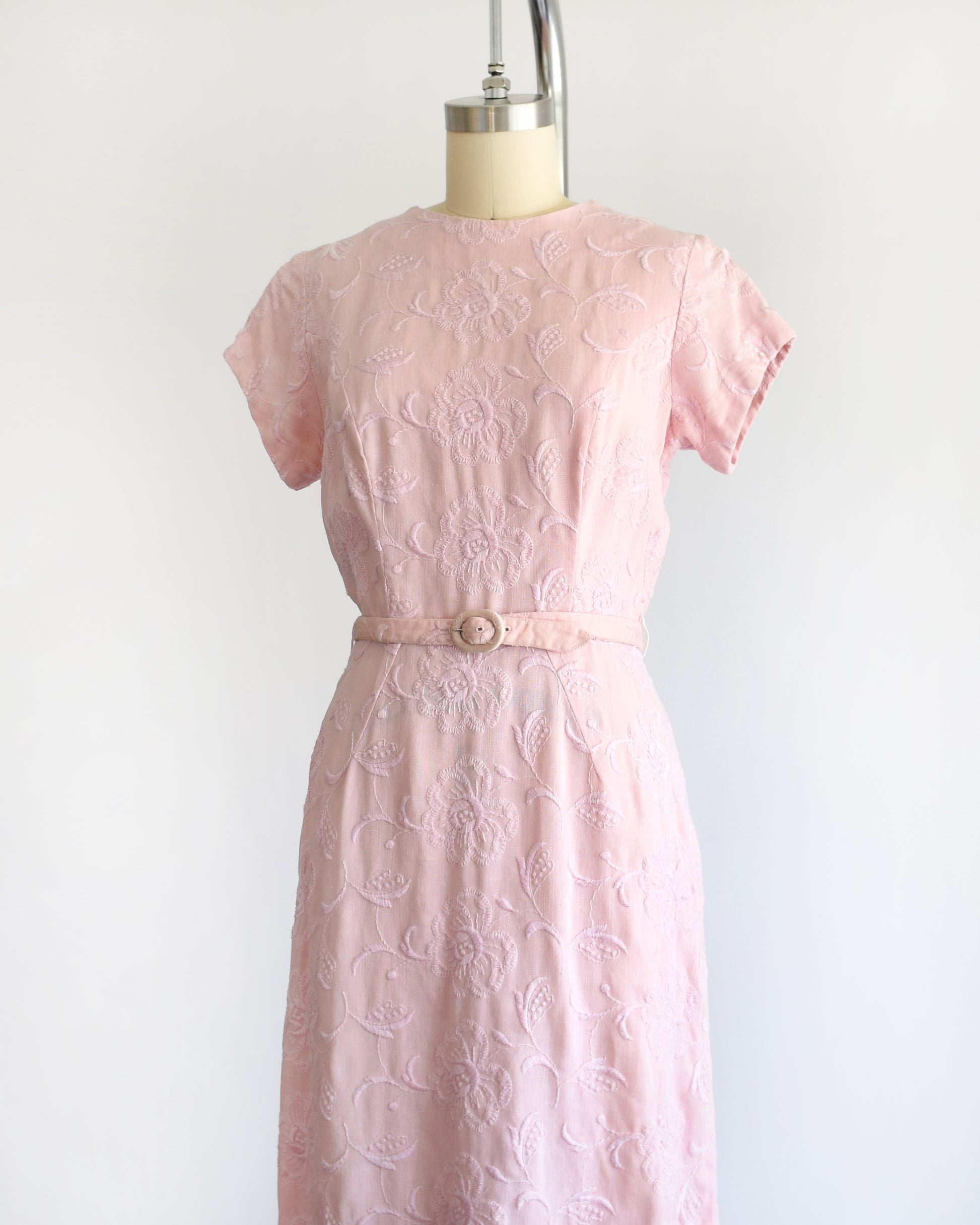 side front view of a vintage 1950s/1960s light pink short-sleeve dress with an embroidered floral pattern and a matching waist belt, displayed on a cream-colored dress form against a white background.