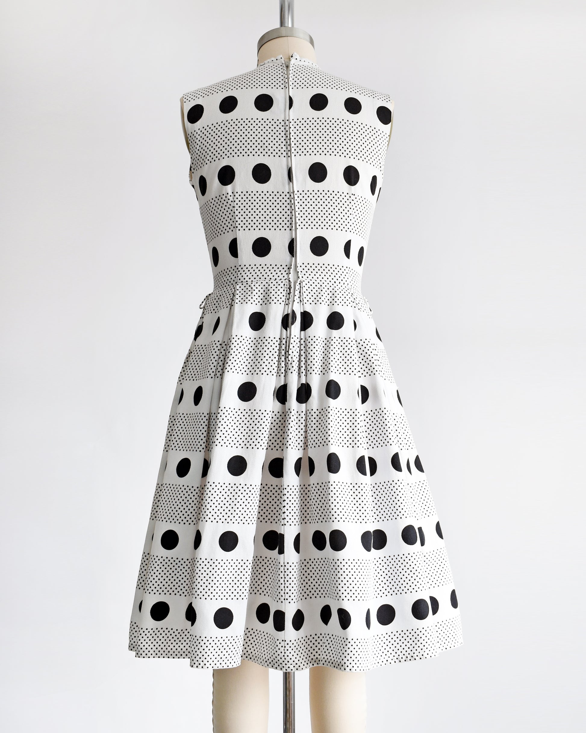 back view of a vintage 1960s polka dot dress that is white cotton piqué with large and small black polka dots in a striped pattern on a dress form