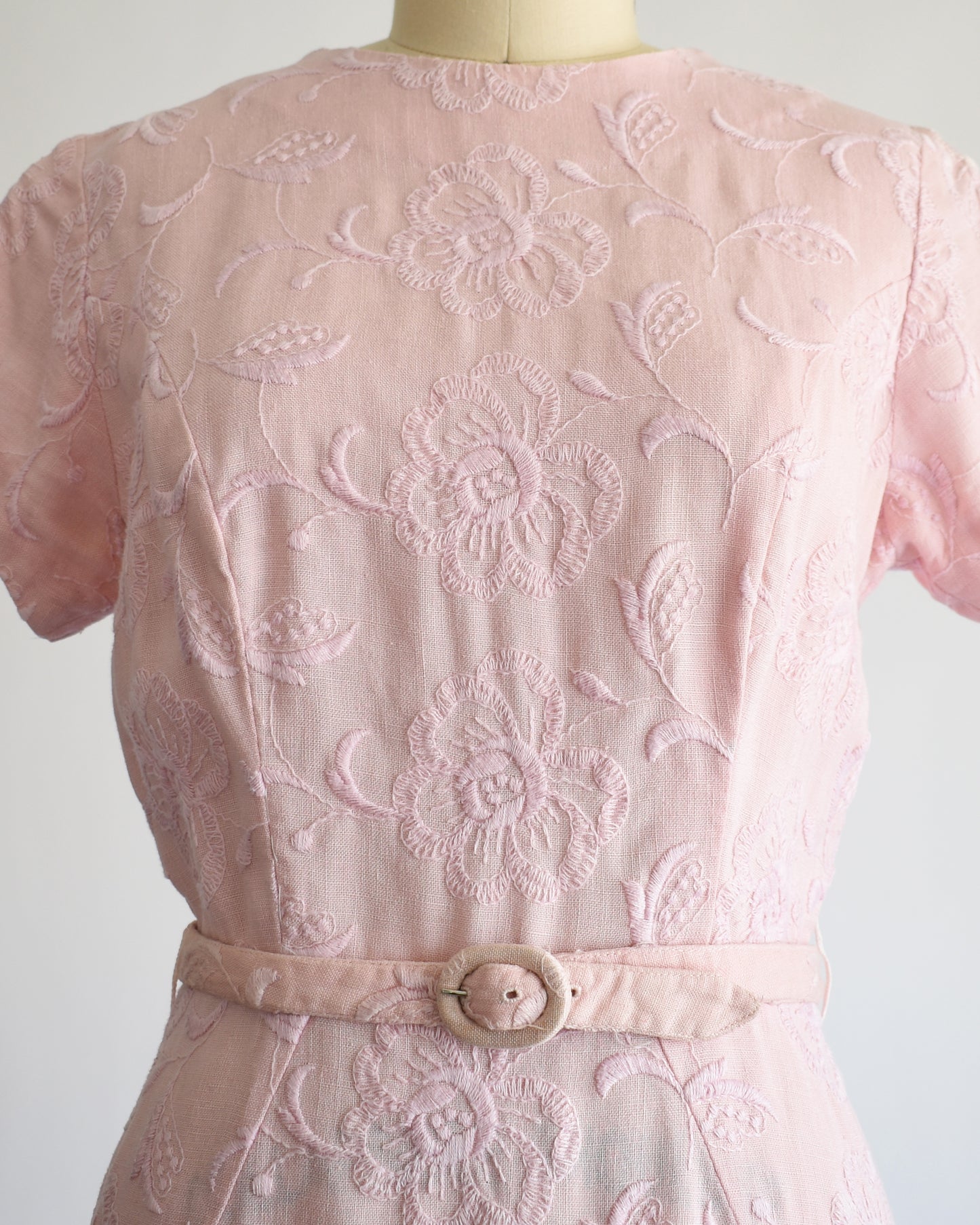 close up of the bodice of aa vintage 1950s/1960s light pink short-sleeve dress with an embroidered floral pattern and a matching waist belt, displayed on a cream-colored dress form against a white background.