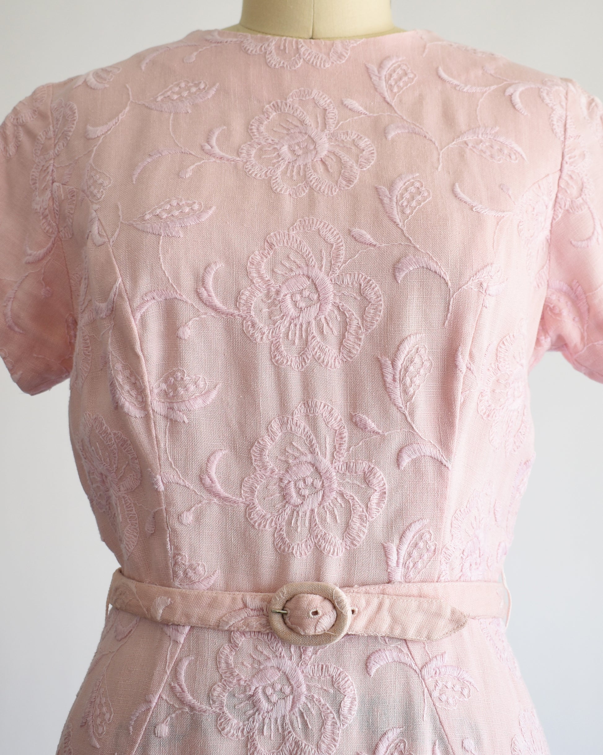 close up of the bodice of aa vintage 1950s/1960s light pink short-sleeve dress with an embroidered floral pattern and a matching waist belt, displayed on a cream-colored dress form against a white background.