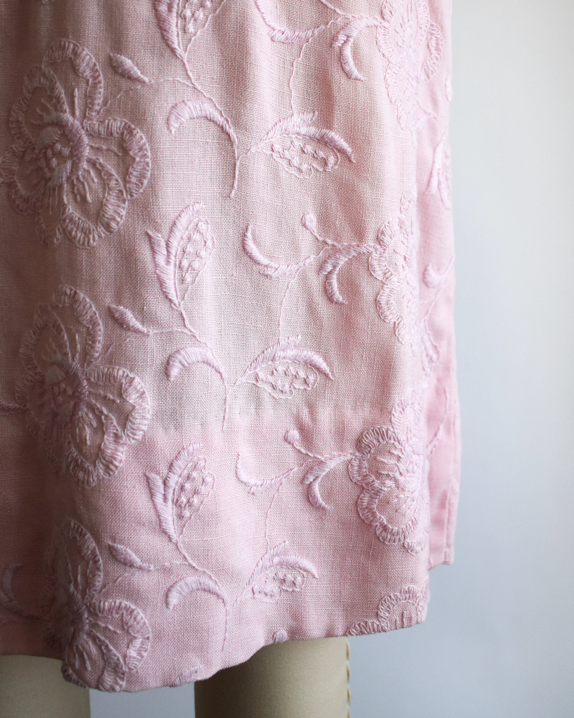 close up of the skirt on a vintage 1950s/1960s light pink short-sleeve dress with an embroidered floral pattern and a matching waist belt, displayed on a cream-colored dress form against a white background.