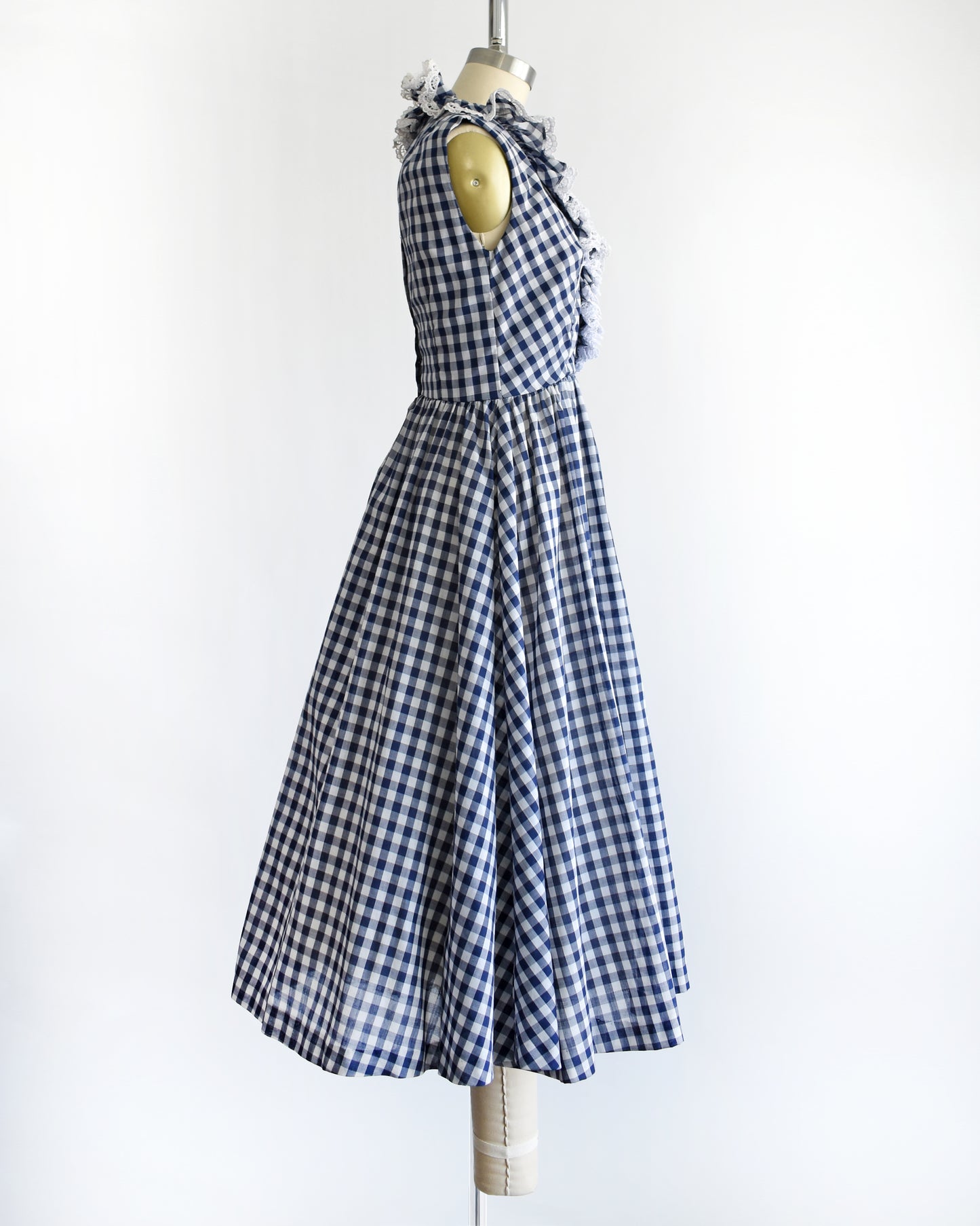 side view of a vintage 1960s sleeveless blue and white gingham dress displayed on a cream dress form against a plain white background.