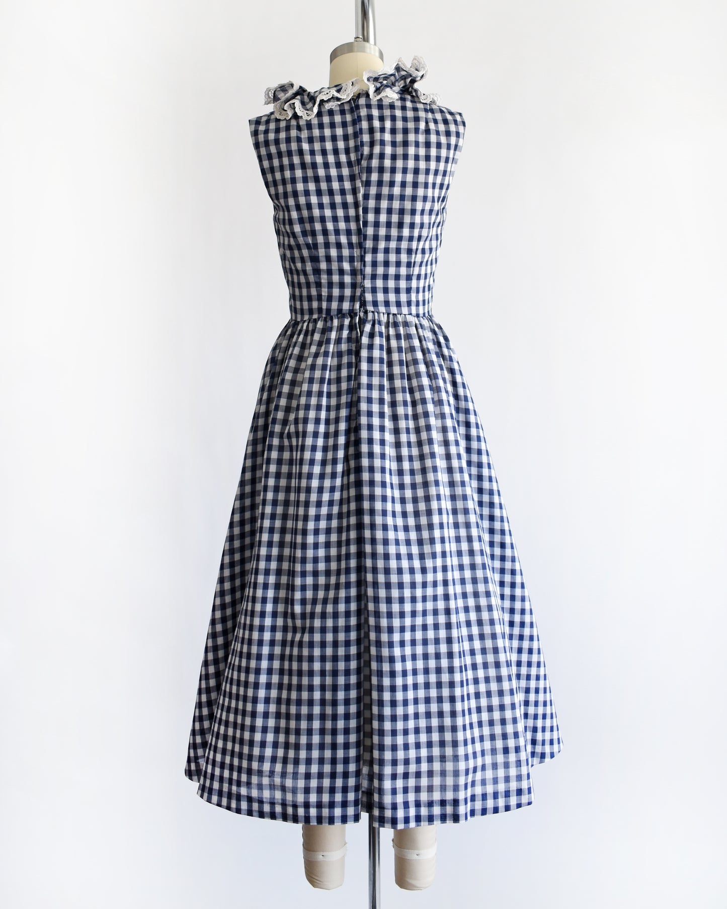 back view of a vintage 1960s sleeveless blue and white gingham dress displayed on a cream dress form against a plain white background.