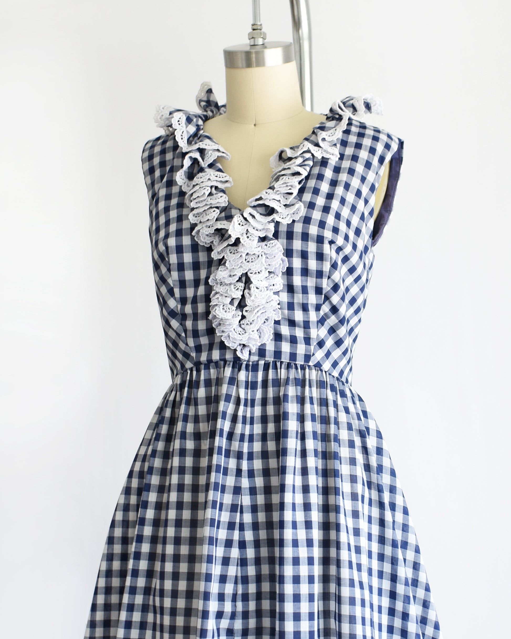 side front view of a vintage 1960s sleeveless blue and white gingham dress displayed on a cream dress form against a plain white background.