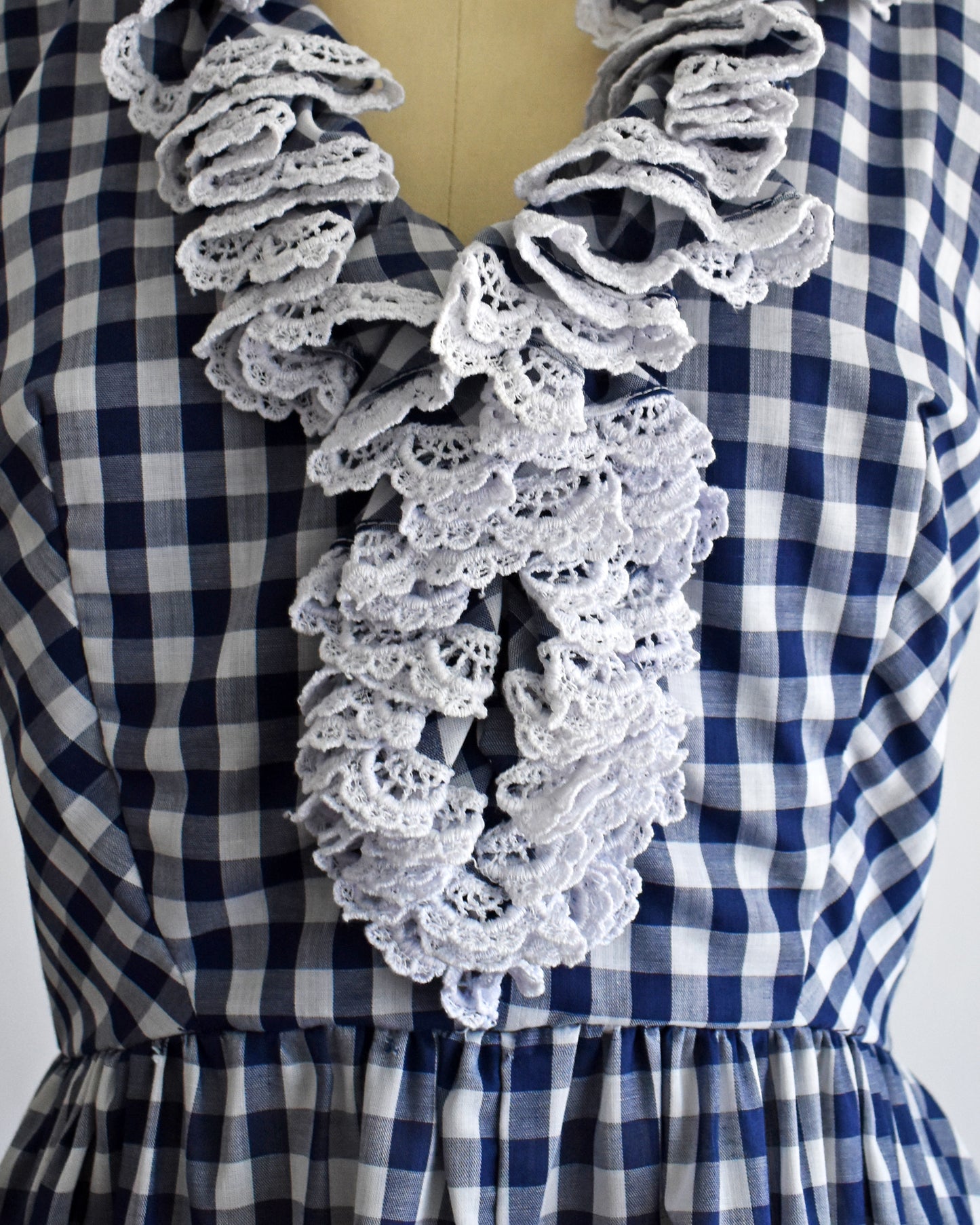 A close-up of a blue and white gingham dress displayed on a mannequin, featuring a large, layered white ruffled collar with intricate crochet lace detailing.