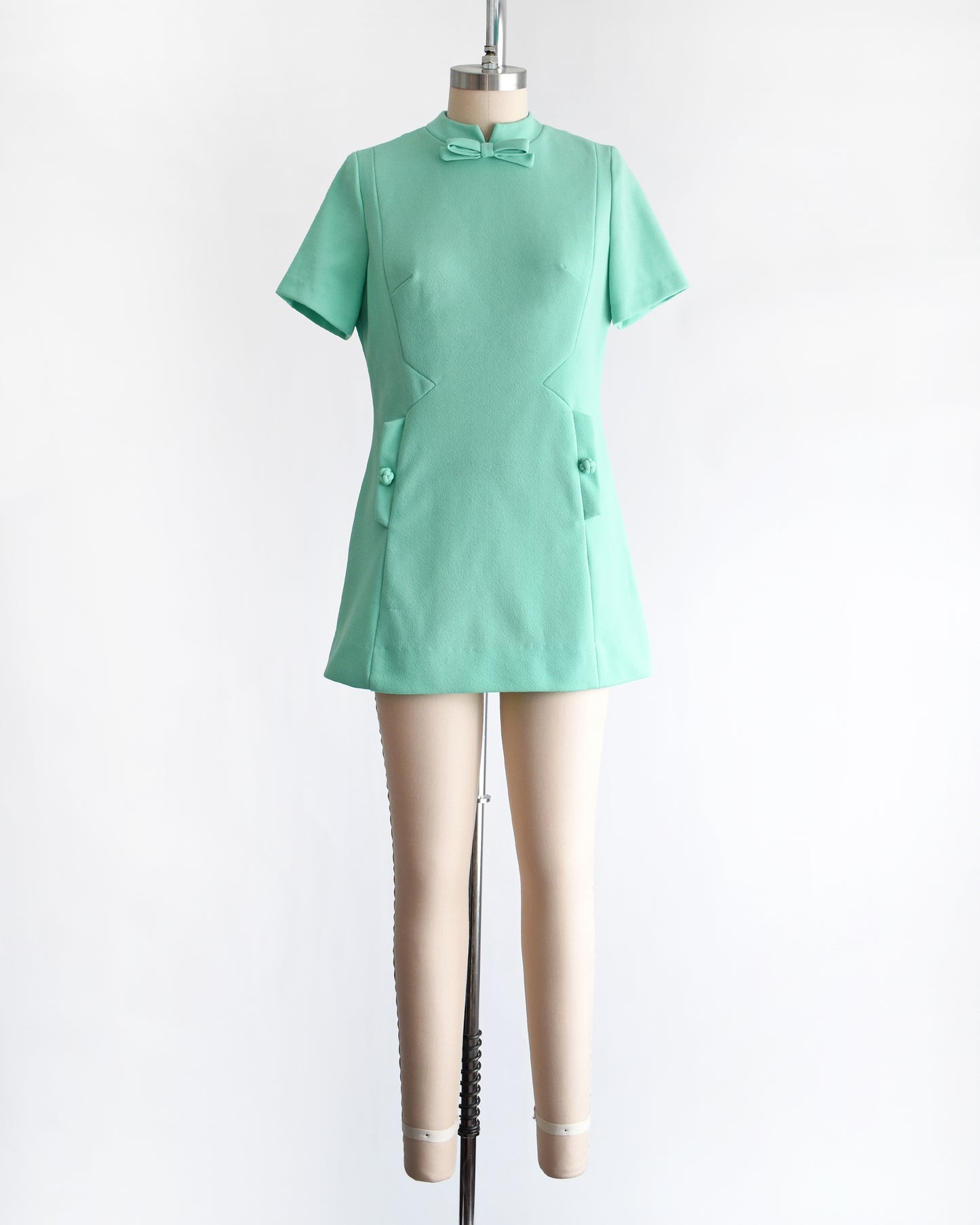 a vintage 1960s aqua green short-sleeve dress with a bow tie neckline, displayed on a beige mannequin against a plain white background.