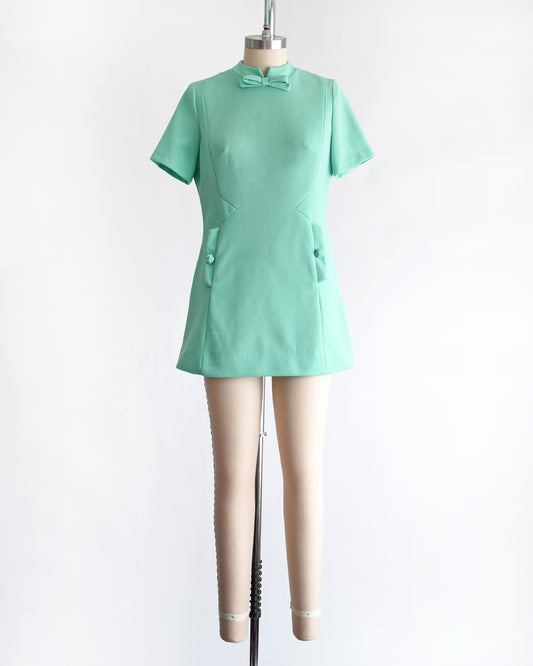 a vintage 1960s aqua green short-sleeve dress with a bow tie neckline, displayed on a beige mannequin against a plain white background.