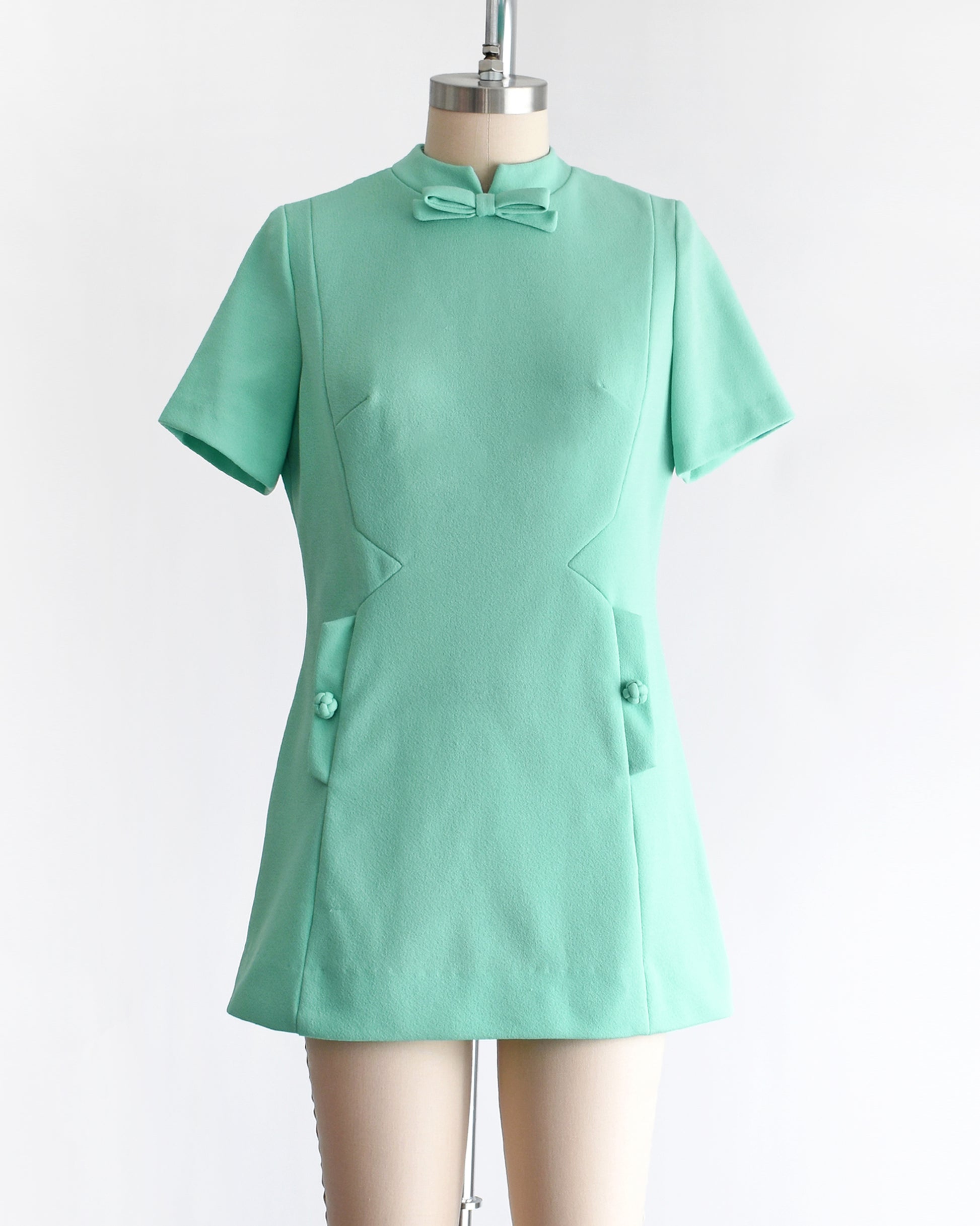 a vintage 1960s aqua green short-sleeve dress with a bow tie neckline, displayed on a beige mannequin against a plain white background.