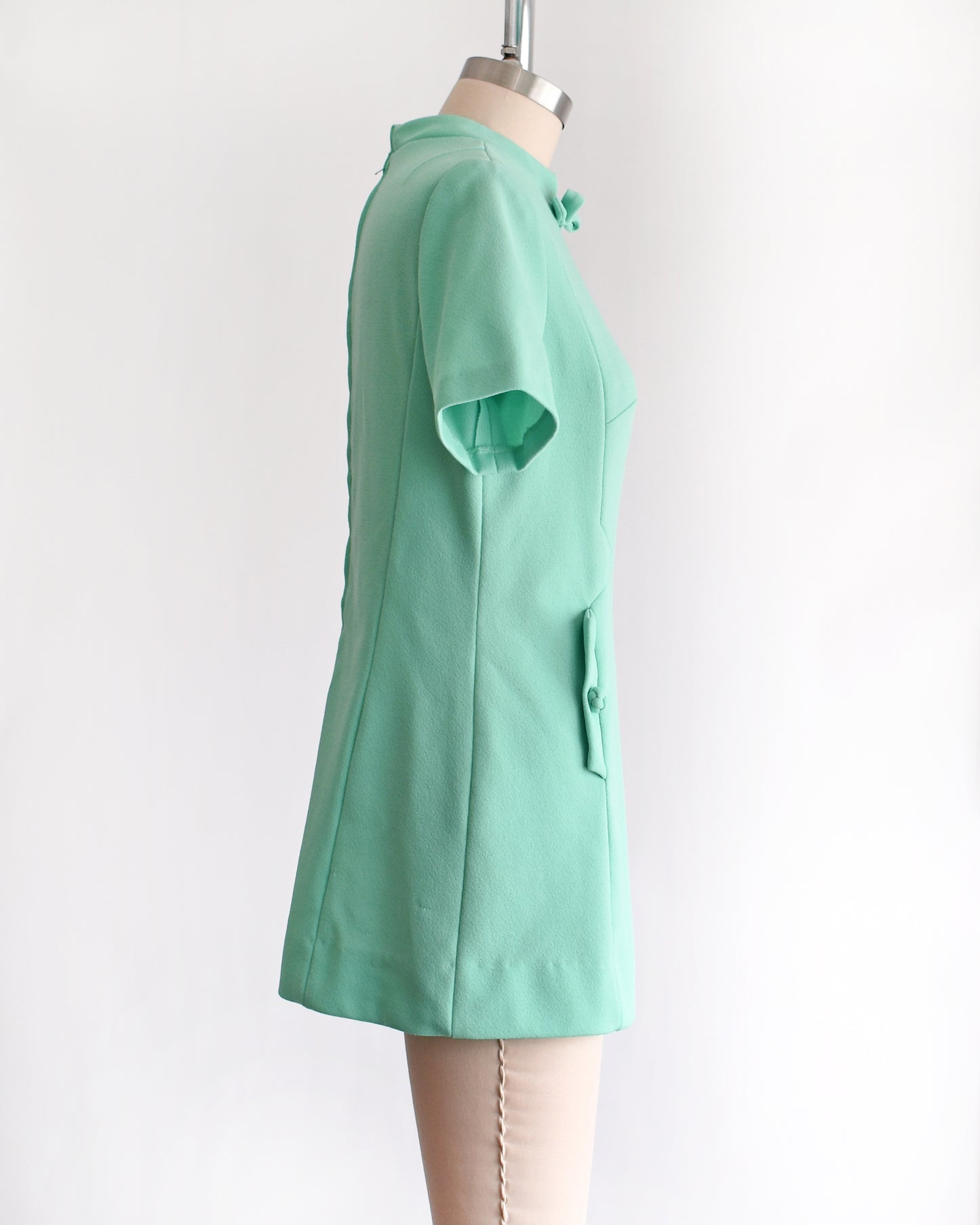 side view of a vintage 1960s aqua green short-sleeve dress with a bow tie neckline, displayed on a beige mannequin against a plain white background.