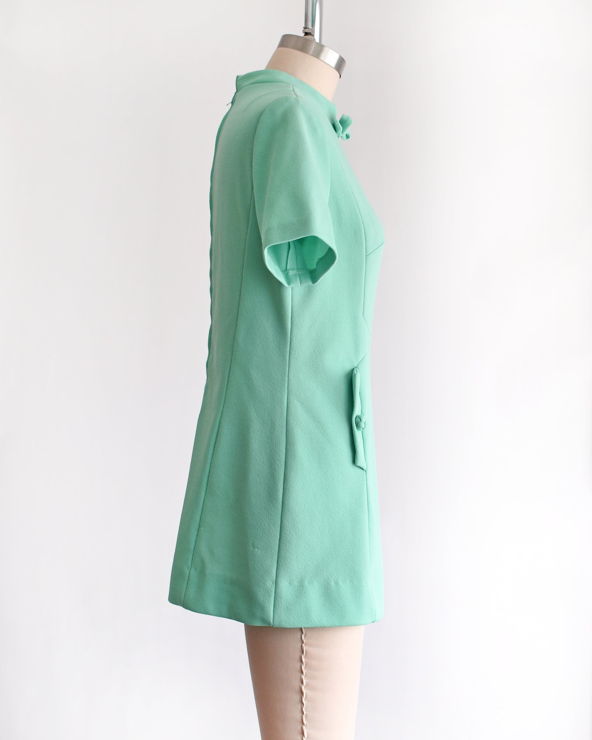 side view of a vintage 1960s aqua green short-sleeve dress with a bow tie neckline, displayed on a beige mannequin against a plain white background.