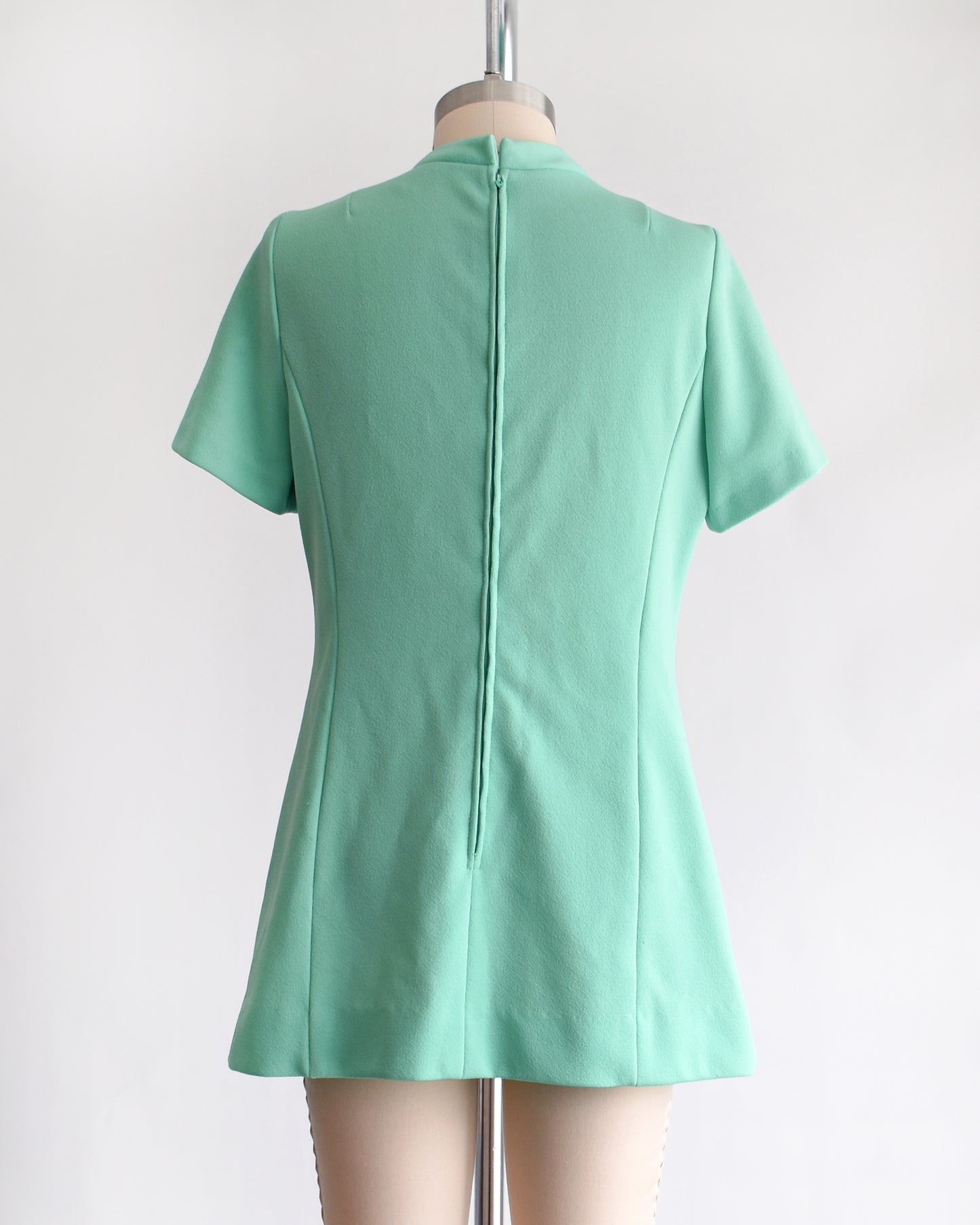 back view of a vintage 1960s aqua green short-sleeve dress with a bow tie neckline, displayed on a beige mannequin against a plain white background.