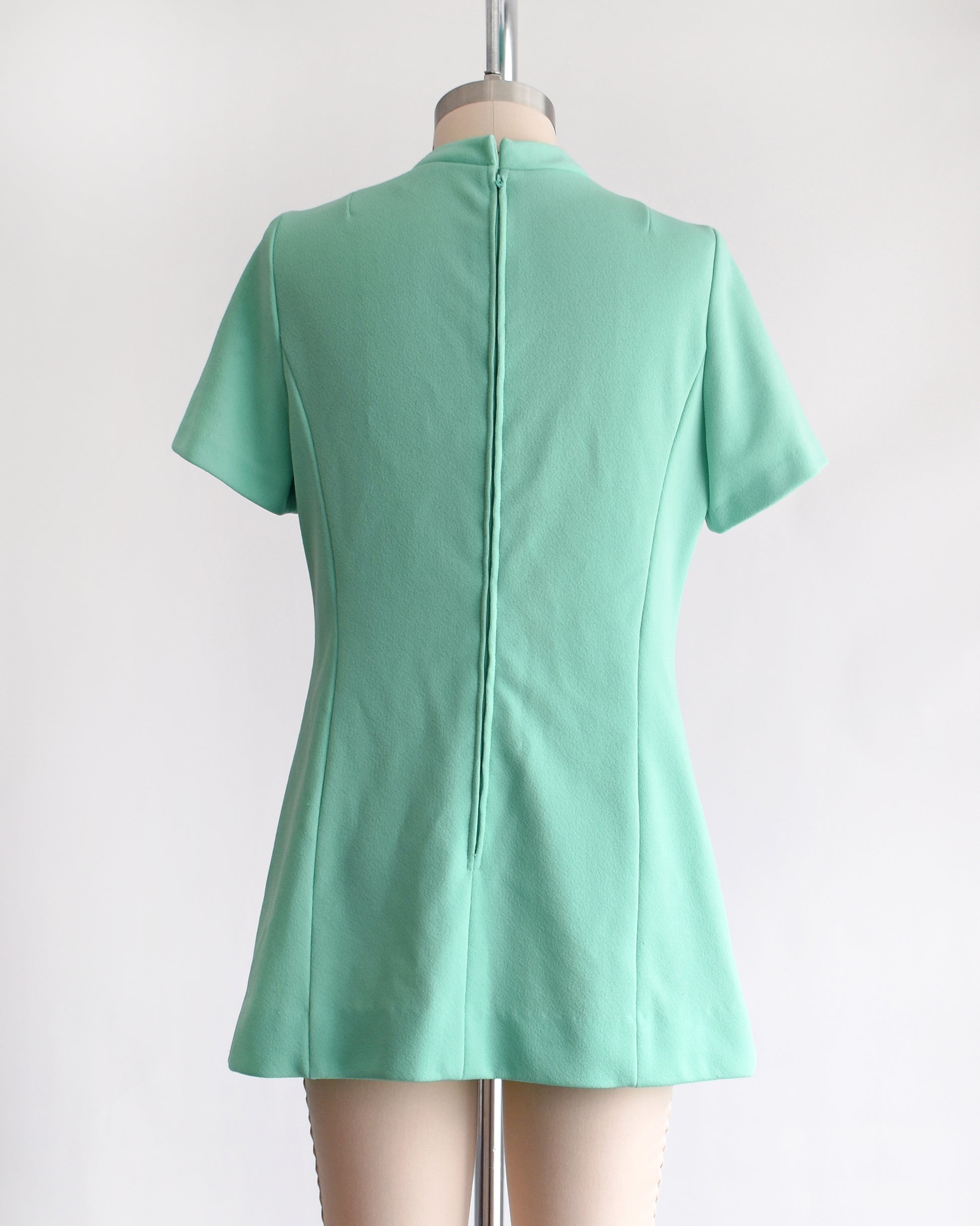 back view of a vintage 1960s aqua green short-sleeve dress with a bow tie neckline, displayed on a beige mannequin against a plain white background.