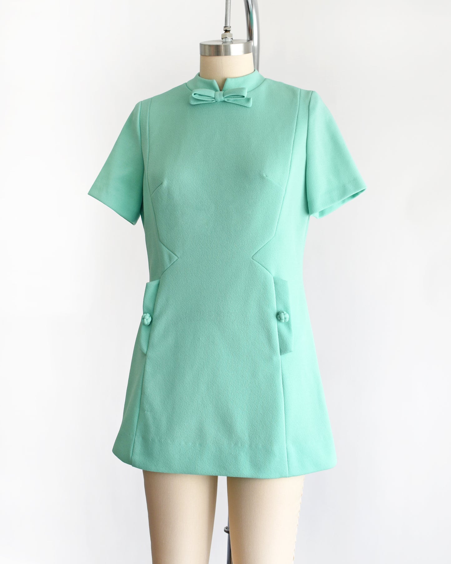 side front view of a vintage 1960s aqua green short-sleeve dress with a bow tie neckline, displayed on a beige mannequin against a plain white background.