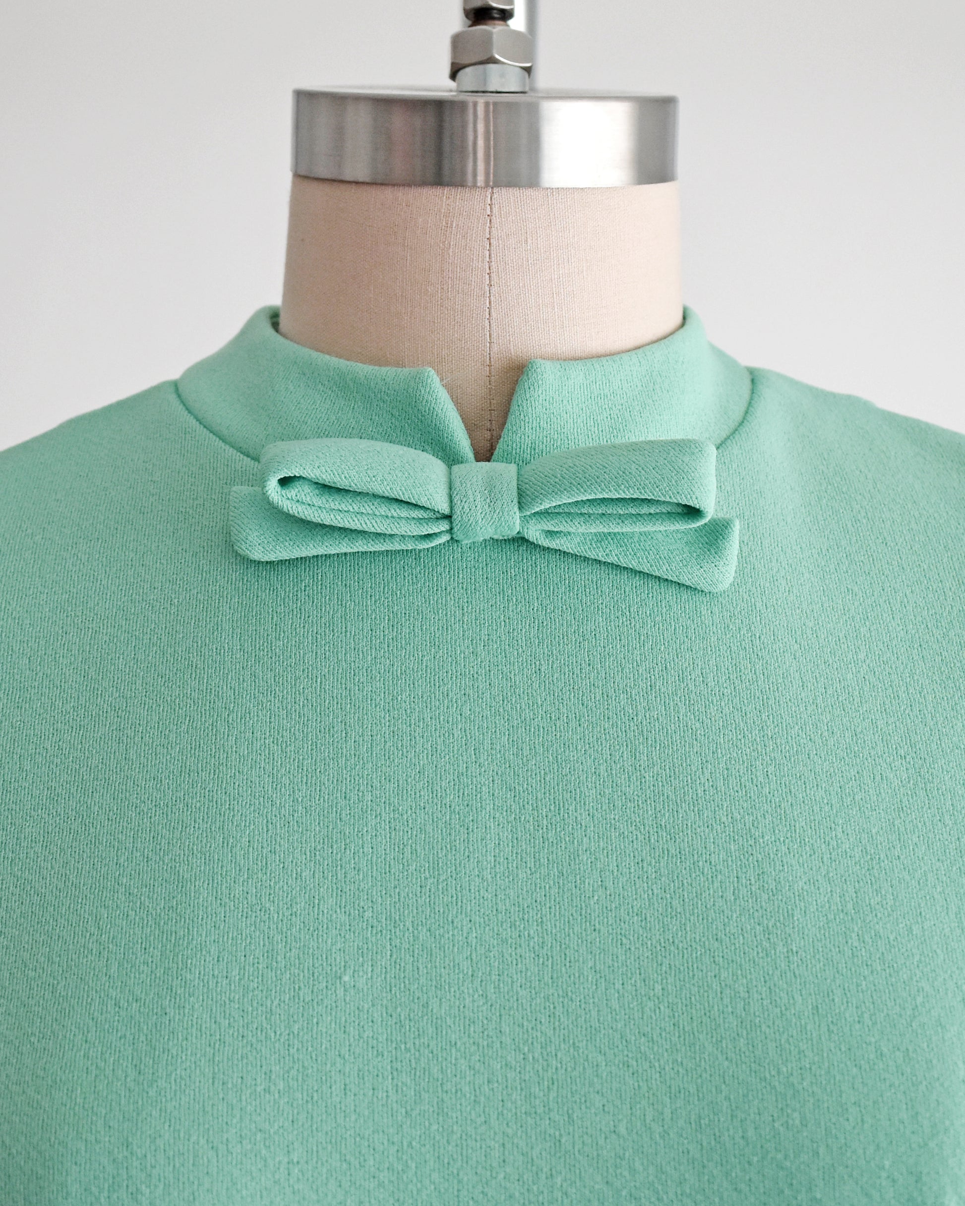 A vintage 1960s aqua green top or dress displayed on a dress form, featuring a high mandarin collar with a matching fabric bow at the neck.