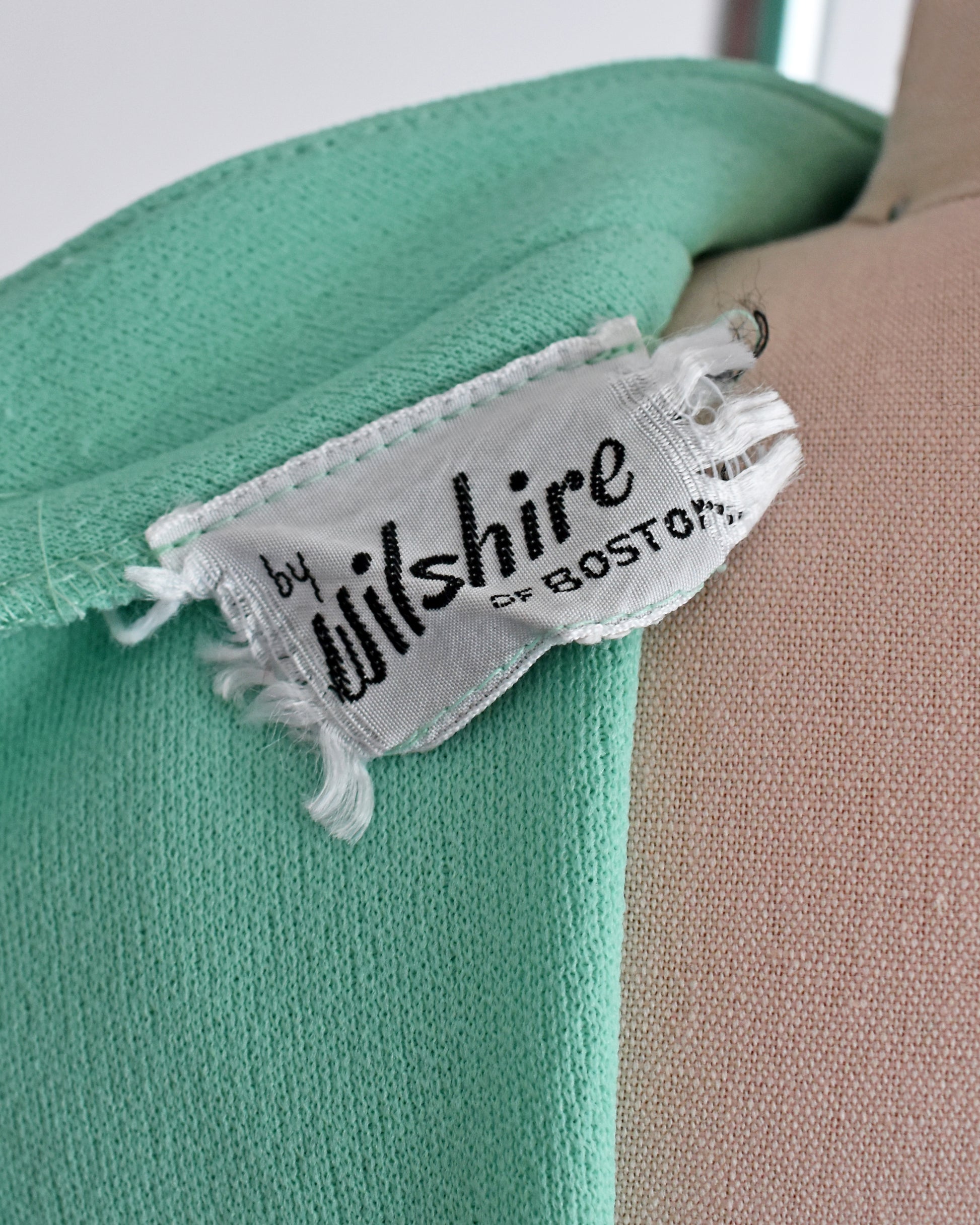 Close-up of a mint green knit garment with a frayed white fabric label reading 'by Wilshire of Boston' on a beige dress form.