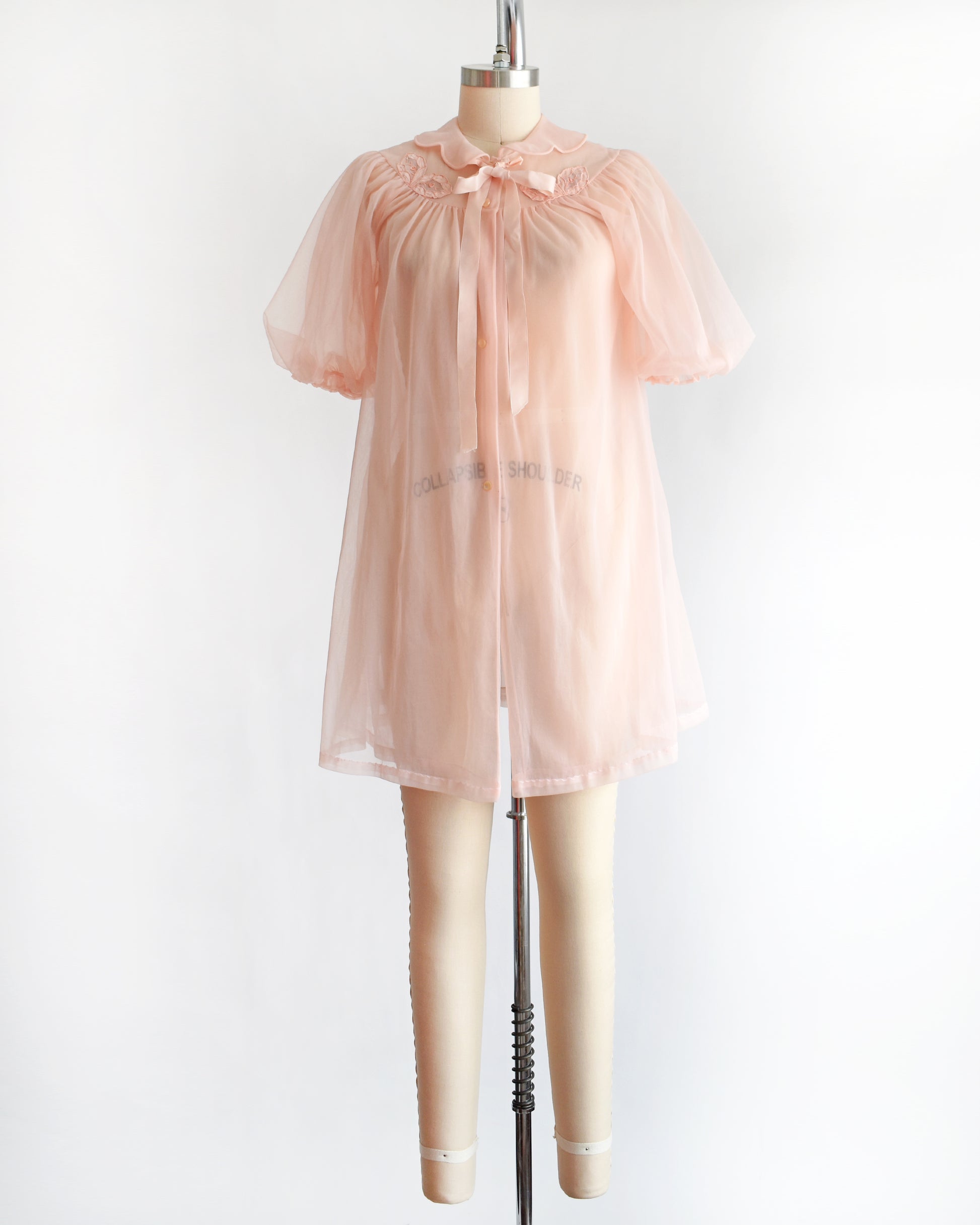 a vintage 1950s/1960s light pink sheer robe with short puff sleeves, a ruffled collar, and a matching bow at the neck, displayed on a mannequin against a plain white background.