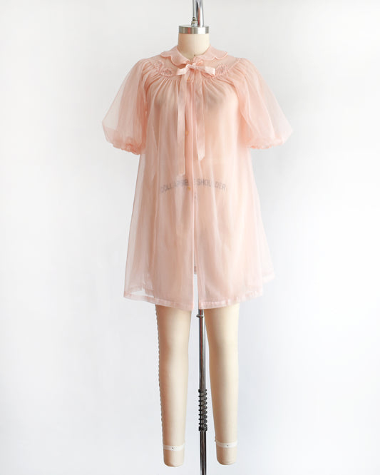 a vintage 1950s/1960s light pink sheer robe with short puff sleeves, a ruffled collar, and a matching bow at the neck, displayed on a mannequin against a plain white background.