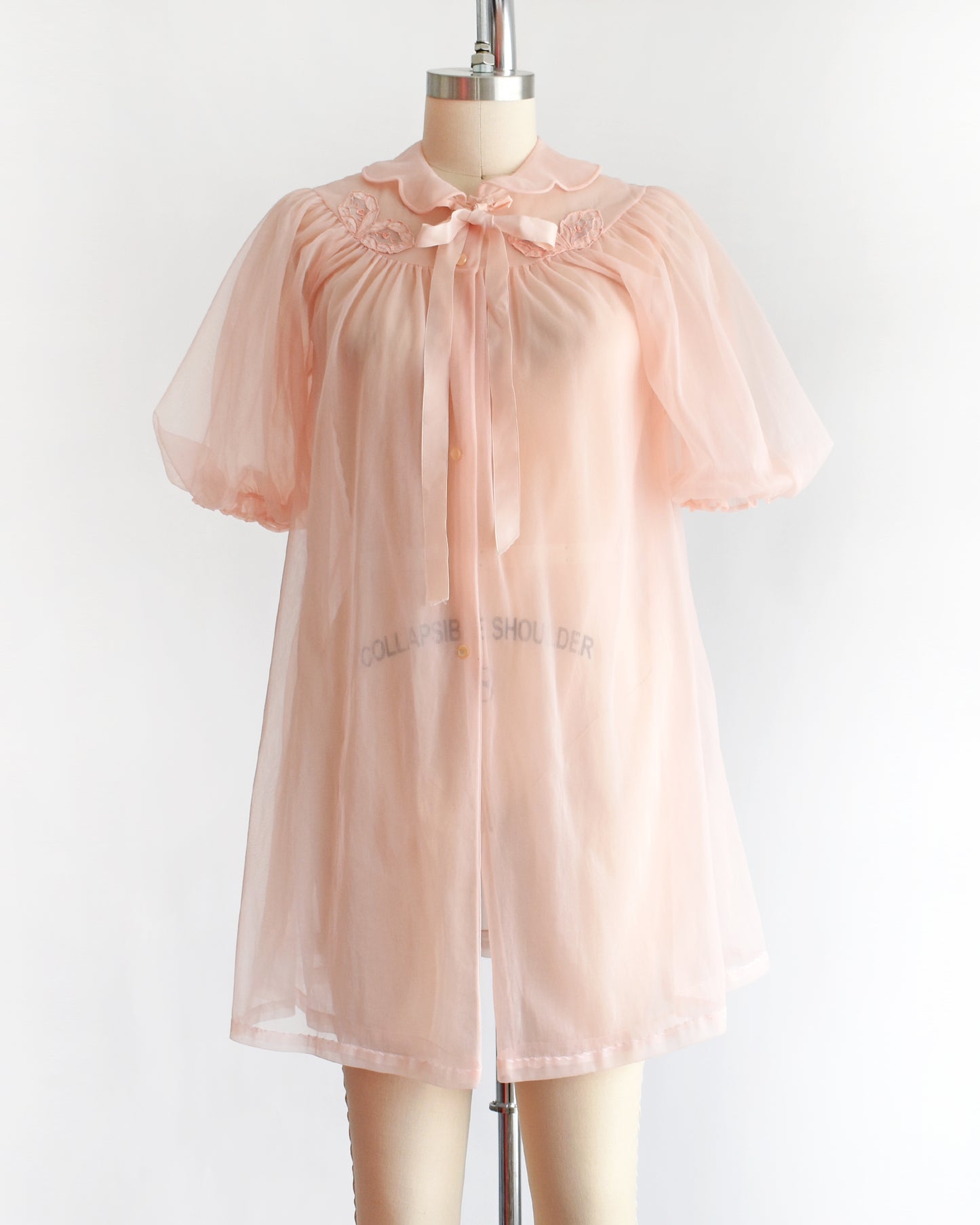 a vintage 1950s/1960s light pink sheer robe with short puff sleeves, a ruffled collar, and a matching bow at the neck, displayed on a mannequin against a plain white background.