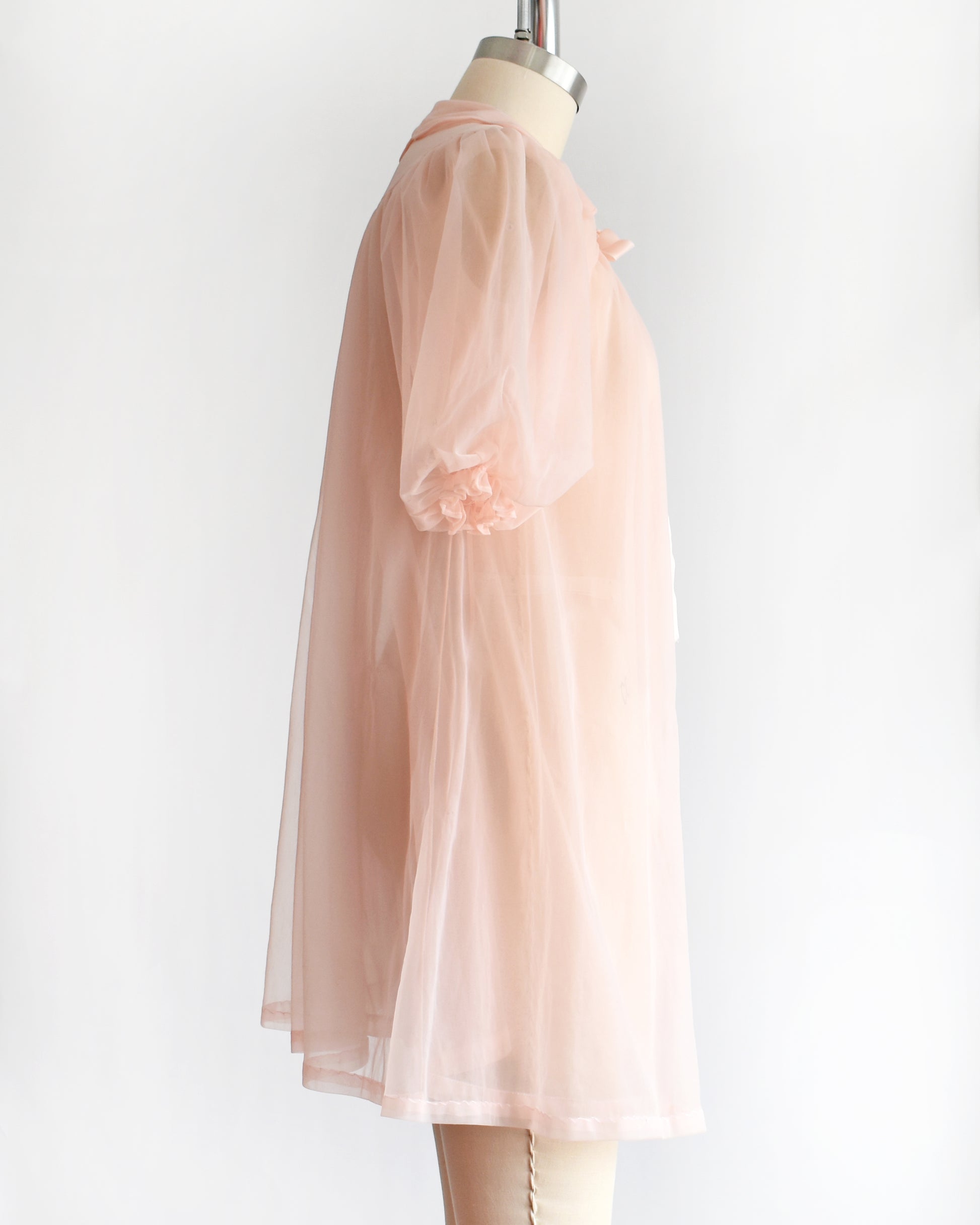 side view of a vintage 1950s/1960s light pink sheer robe with short puff sleeves, a ruffled collar, and a matching bow at the neck, displayed on a mannequin against a plain white background.