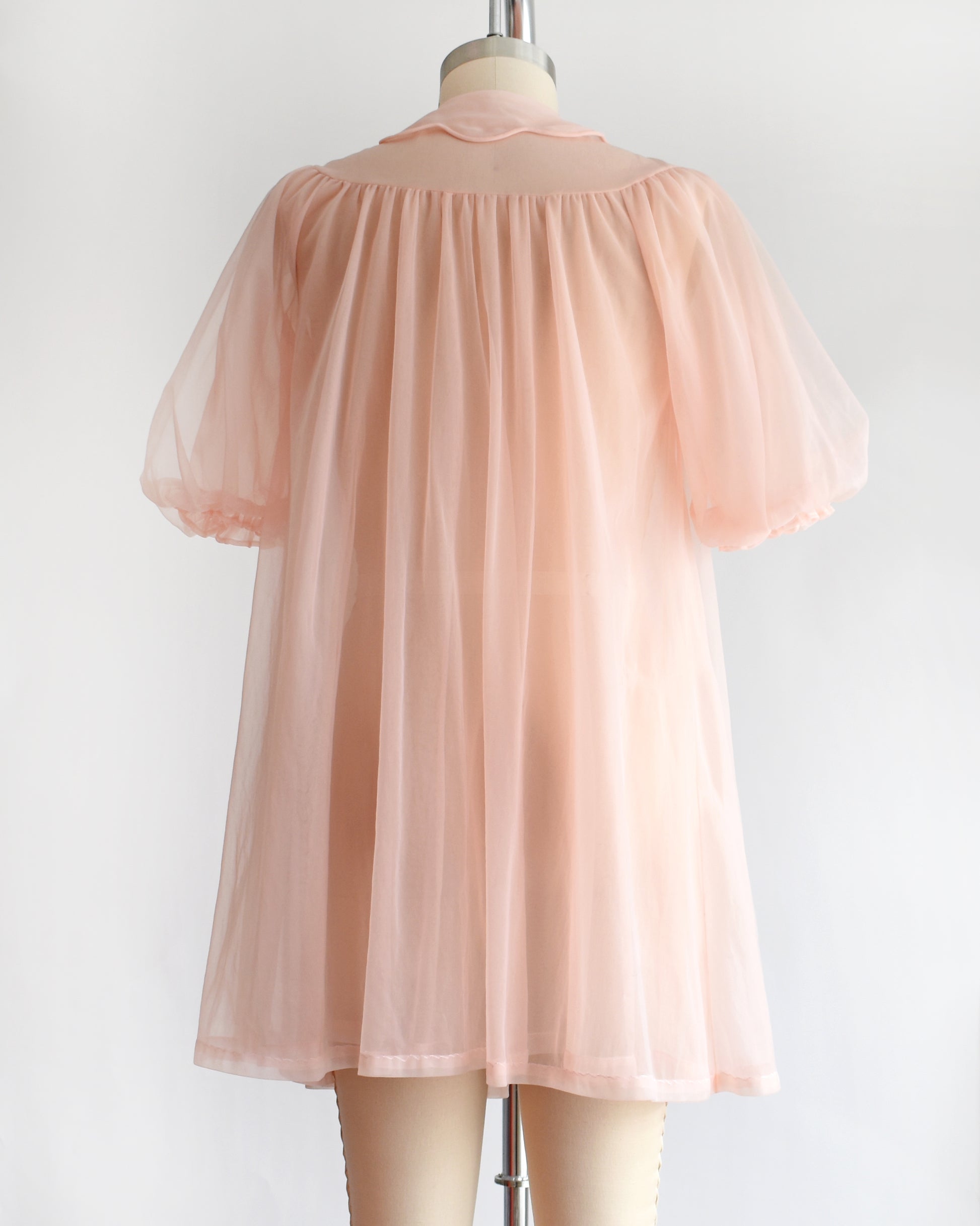 back view of a vintage 1950s/1960s light pink sheer robe with short puff sleeves, a ruffled collar, and a matching bow at the neck, displayed on a mannequin against a plain white background.