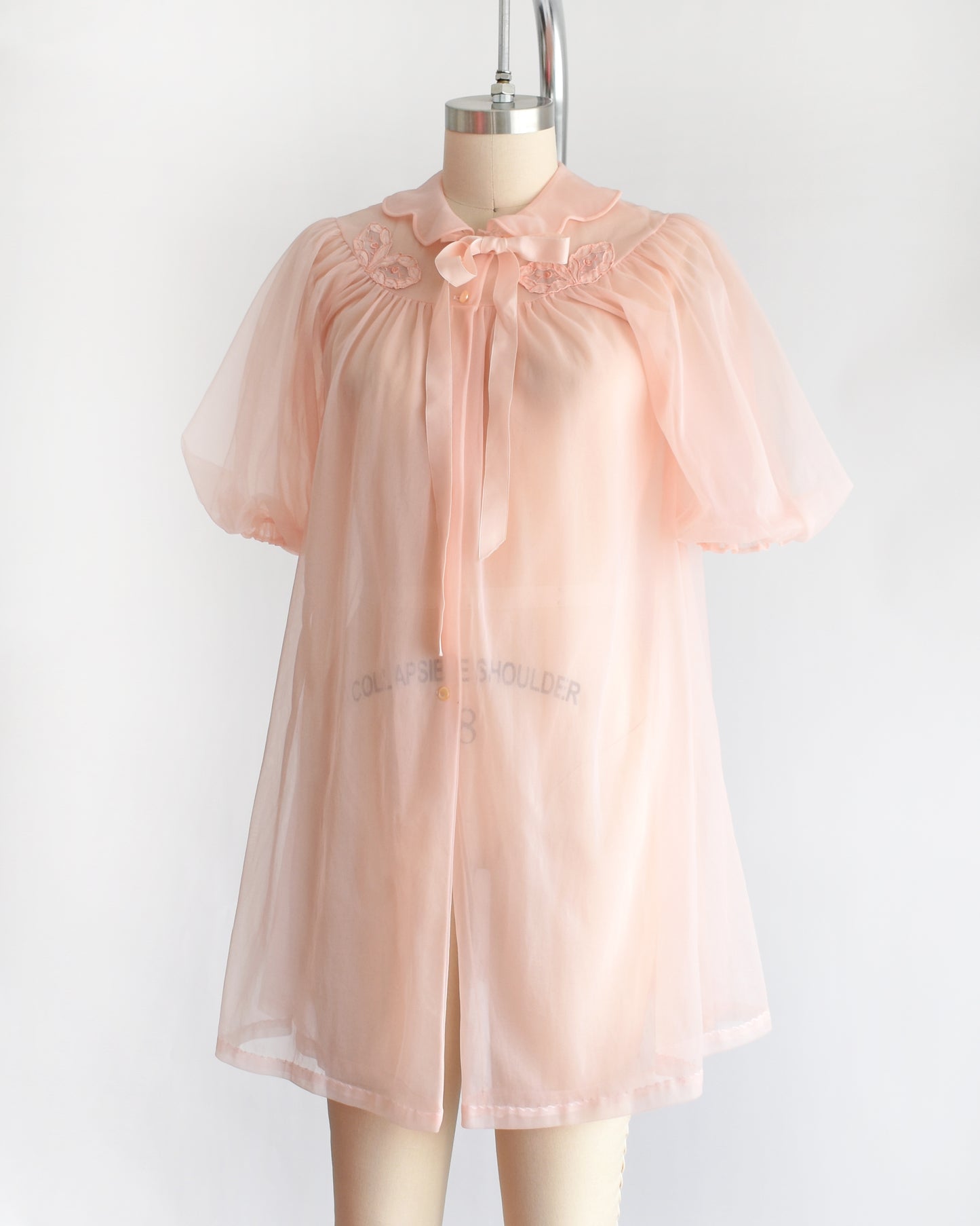 side front view of a vintage 1950s/1960s light pink sheer robe with short puff sleeves, a ruffled collar, and a matching bow at the neck, displayed on a mannequin against a plain white background.
