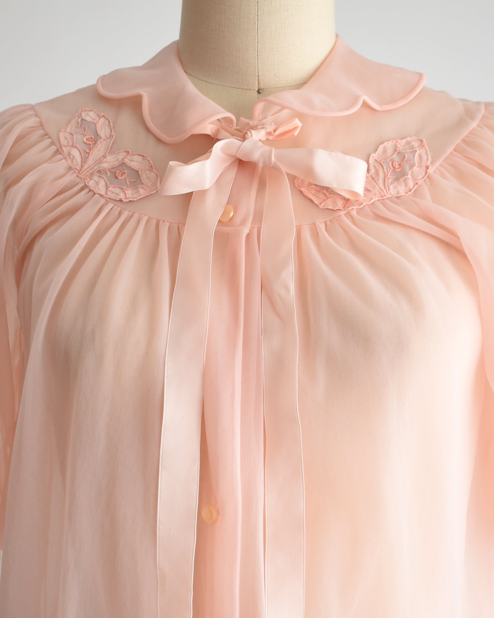 A close-up of a light pink robe displayed on a mannequin. The blouse features a ruffled Peter Pan collar, a matching satin bow at the neck, and embroidered floral details on the shoulders.
