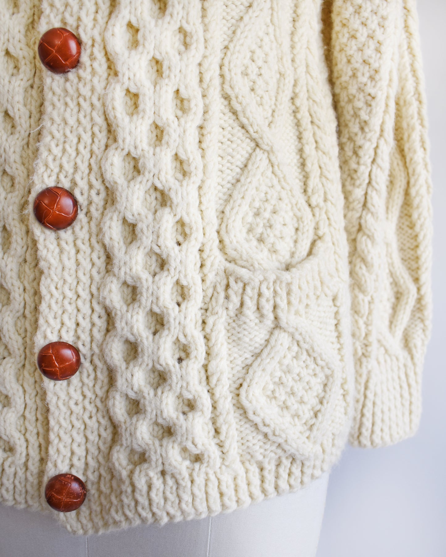 close up of the buttons and pocket on a vintage 1970s/1980s cream-colored cable knit cardigan on a mannequin against a white background