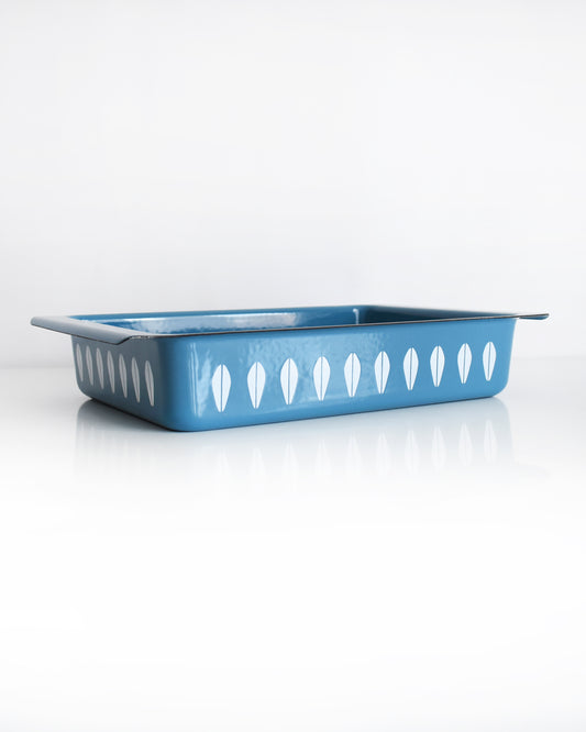 A mid century modern blue Cathrineholm lasagna pan that is rectangular with a decorative pattern of white lotus shapes on the sides, shown on a clean white background with a soft reflection.