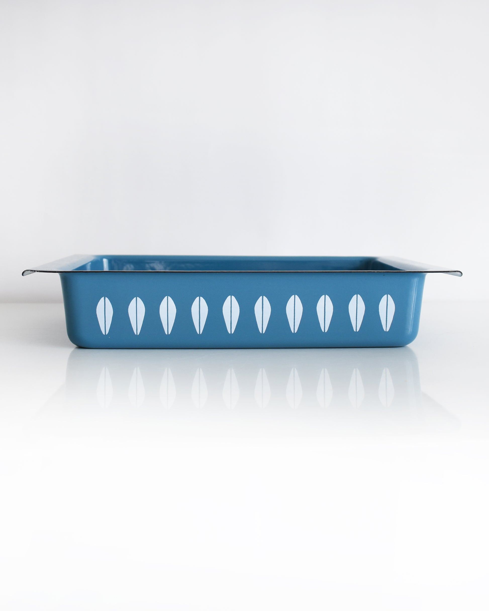 A mid century modern blue Cathrineholm lasagna pan that is rectangular with a decorative pattern of white lotus shapes on the sides, shown on a clean white background with a soft reflection.