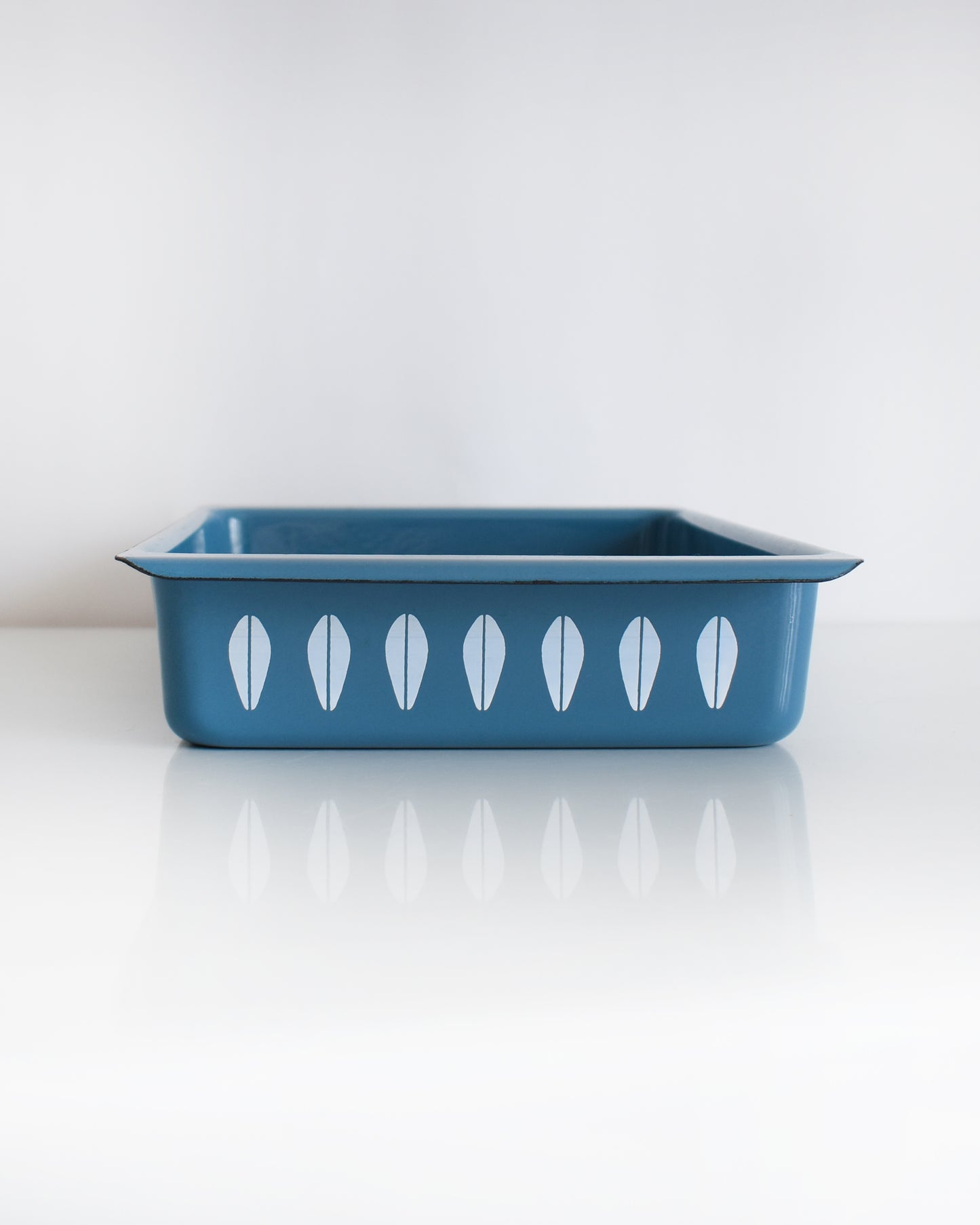 A mid century modern blue Cathrineholm lasagna pan that is rectangular with a decorative pattern of white lotus shapes on the sides, shown on a clean white background with a soft reflection.