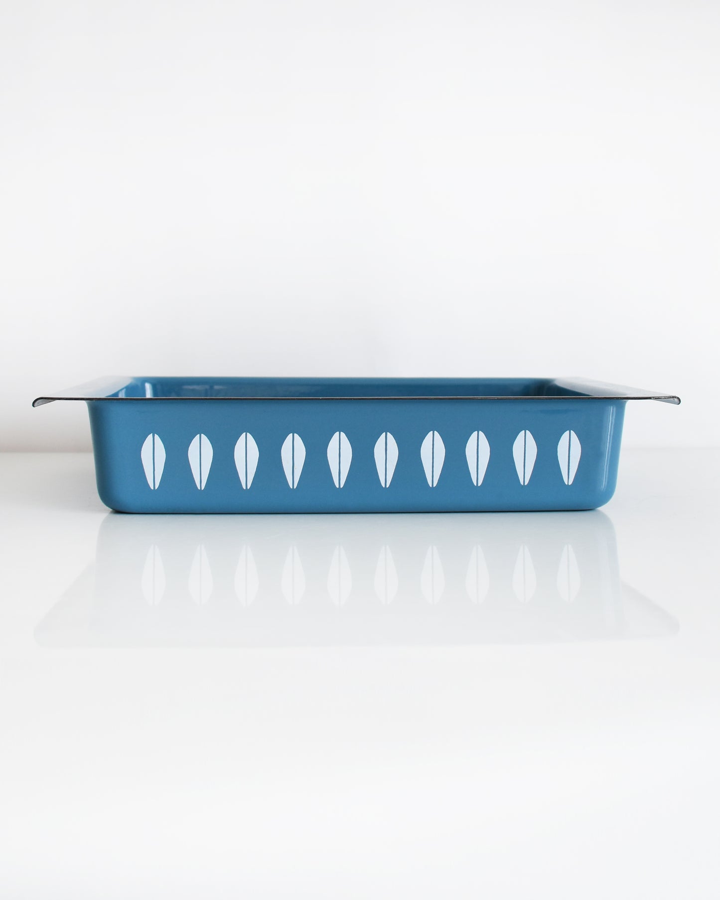 A mid century modern blue Cathrineholm lasagna pan that is rectangular with a decorative pattern of white lotus shapes on the sides, shown on a clean white background with a soft reflection.