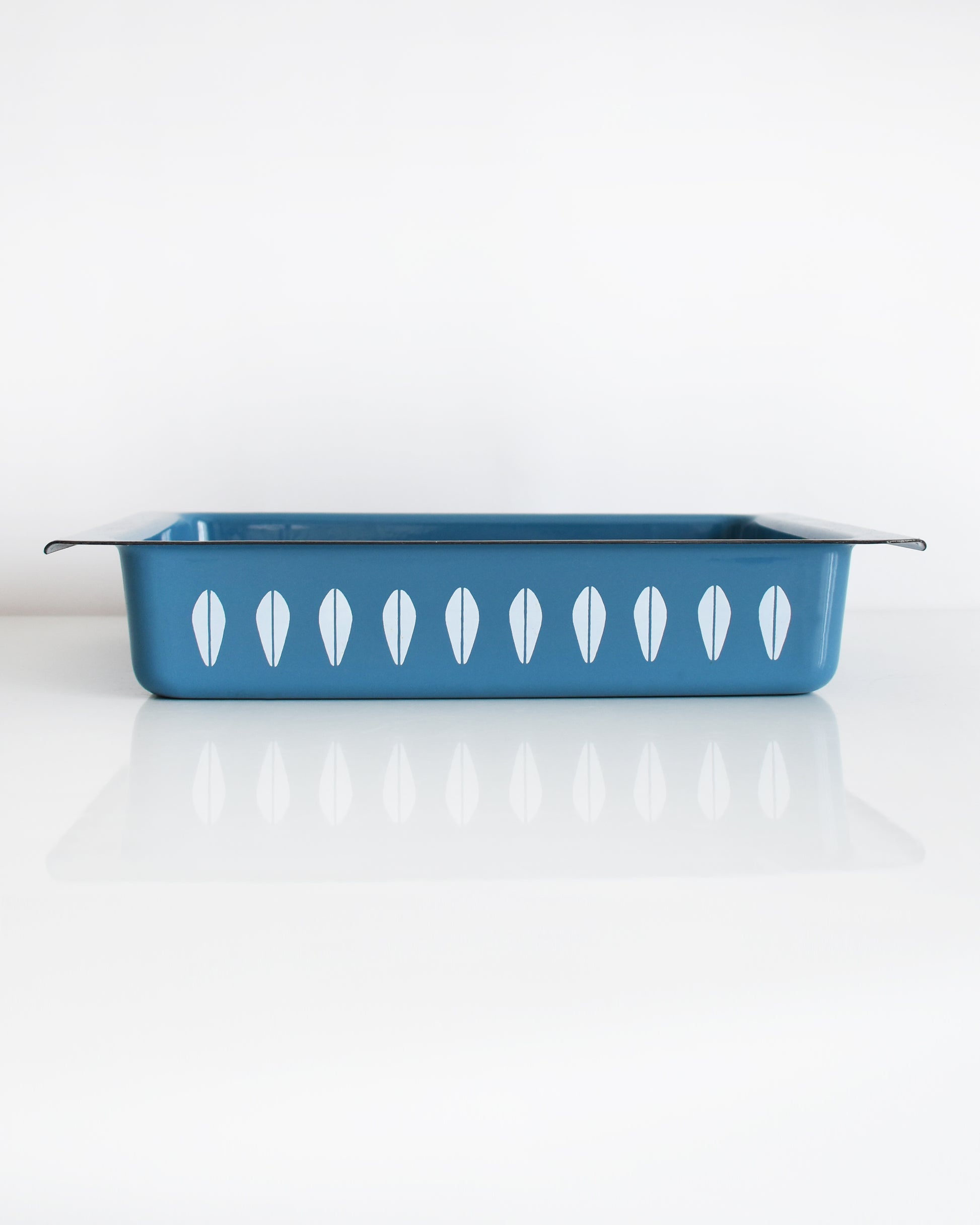 A mid century modern blue Cathrineholm lasagna pan that is rectangular with a decorative pattern of white lotus shapes on the sides, shown on a clean white background with a soft reflection.