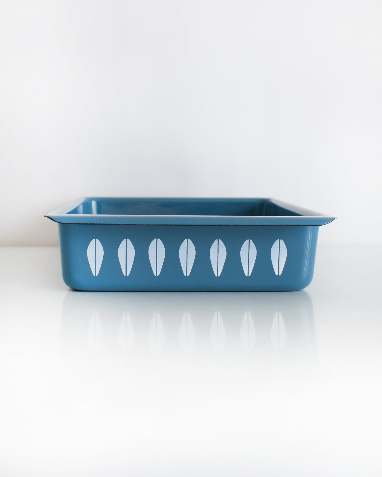 A mid century modern blue Cathrineholm lasagna pan that is rectangular with a decorative pattern of white lotus shapes on the sides, shown on a clean white background with a soft reflection.