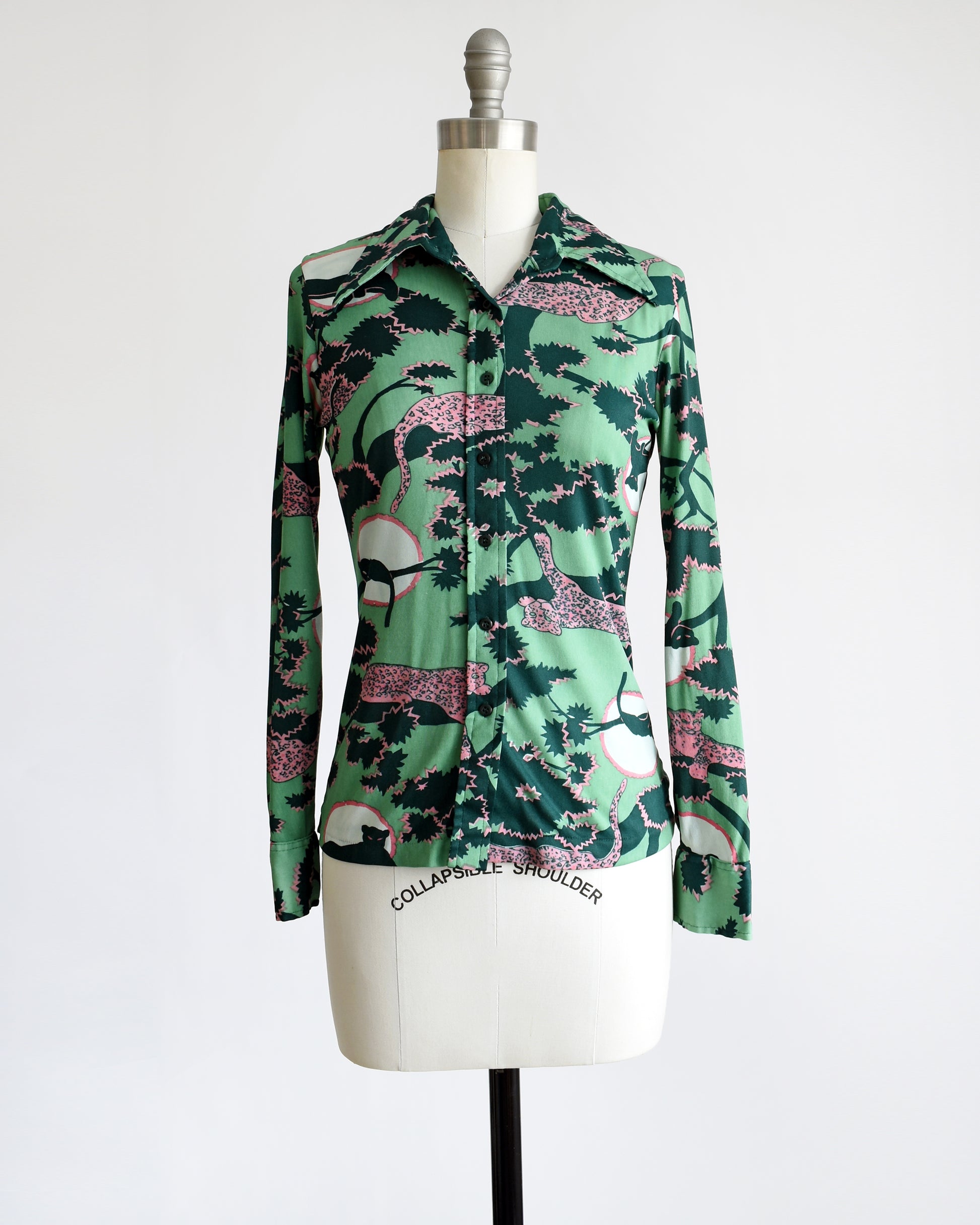 a vintage 1970s disco blouse that is green and a print of pink and black leopards lounging in dark green and pink trees. The black leopards are set against a white circle with pink trim.