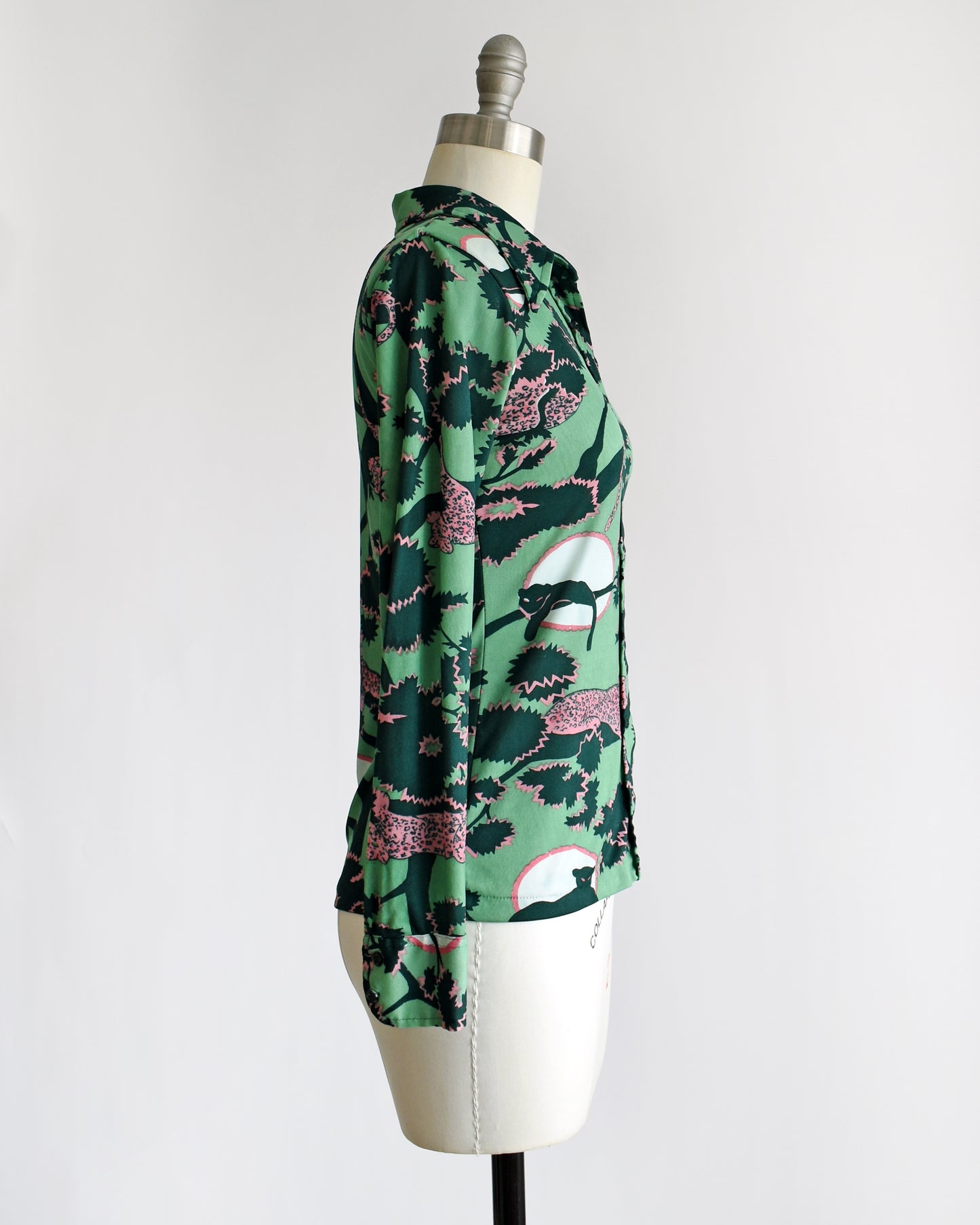 side view of a vintage 1970s disco blouse that is green and a print of pink and black leopards lounging in dark green and pink trees. The black leopards are set against a white circle with pink trim.