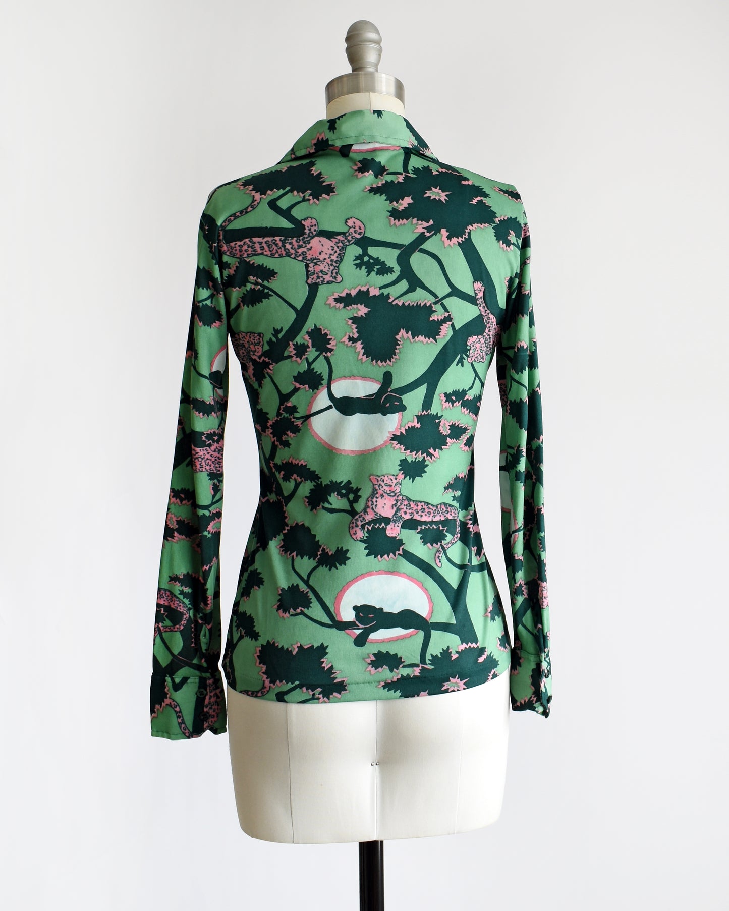 back view of a vintage 1970s disco blouse that is green and a print of pink and black leopards lounging in dark green and pink trees. The black leopards are set against a white circle with pink trim.