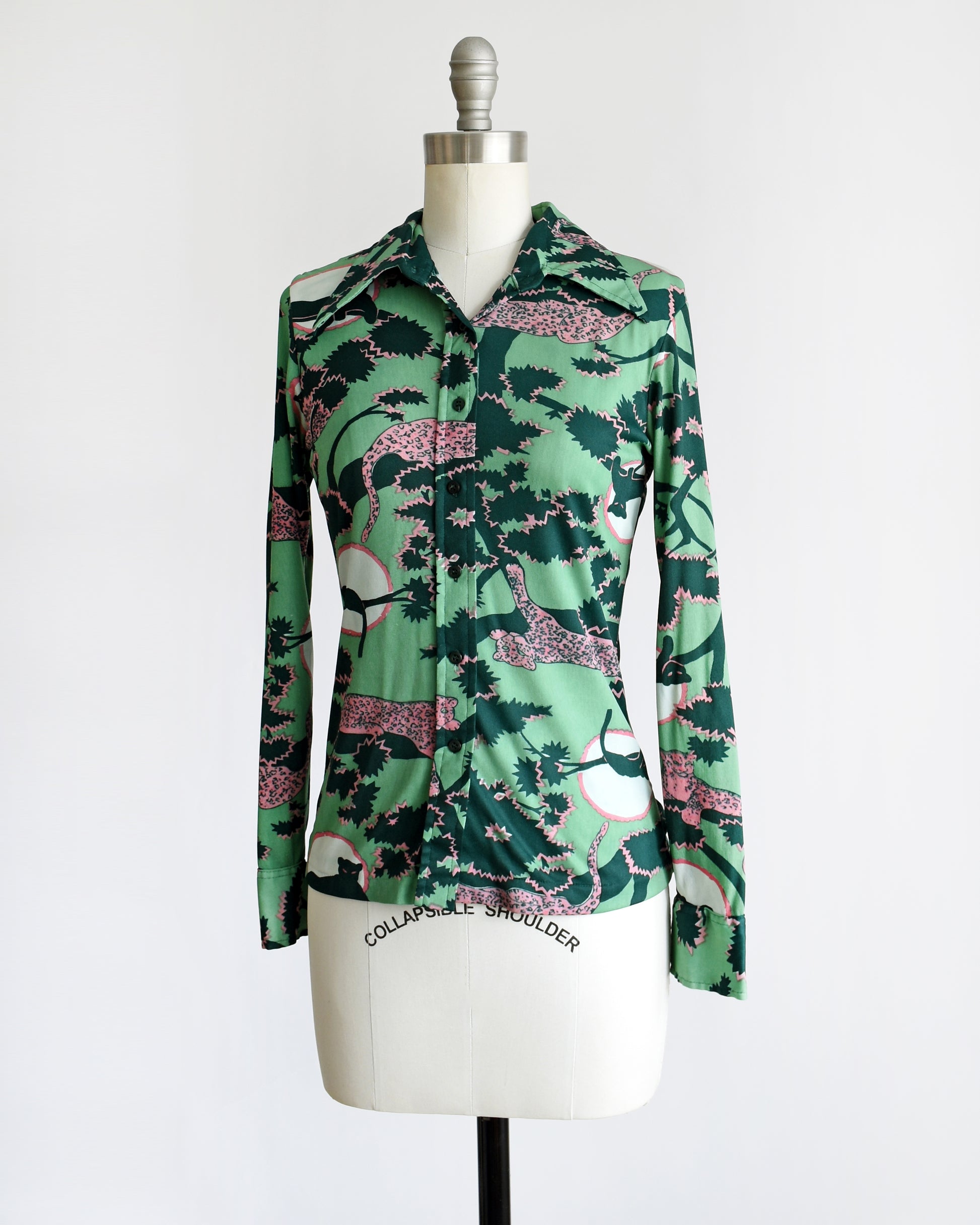 front side view of a vintage 1970s disco blouse that is green and a print of pink and black leopards lounging in dark green and pink trees. The black leopards are set against a white circle with pink trim.