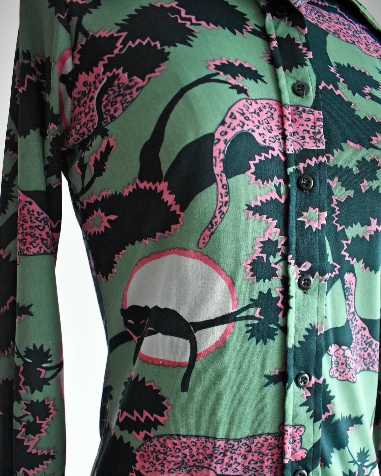 close up of the  print of a vintage 1970s disco blouse that is green and a print of pink and black leopards lounging in dark green and pink trees. The black leopards are set against a white circle with pink trim.