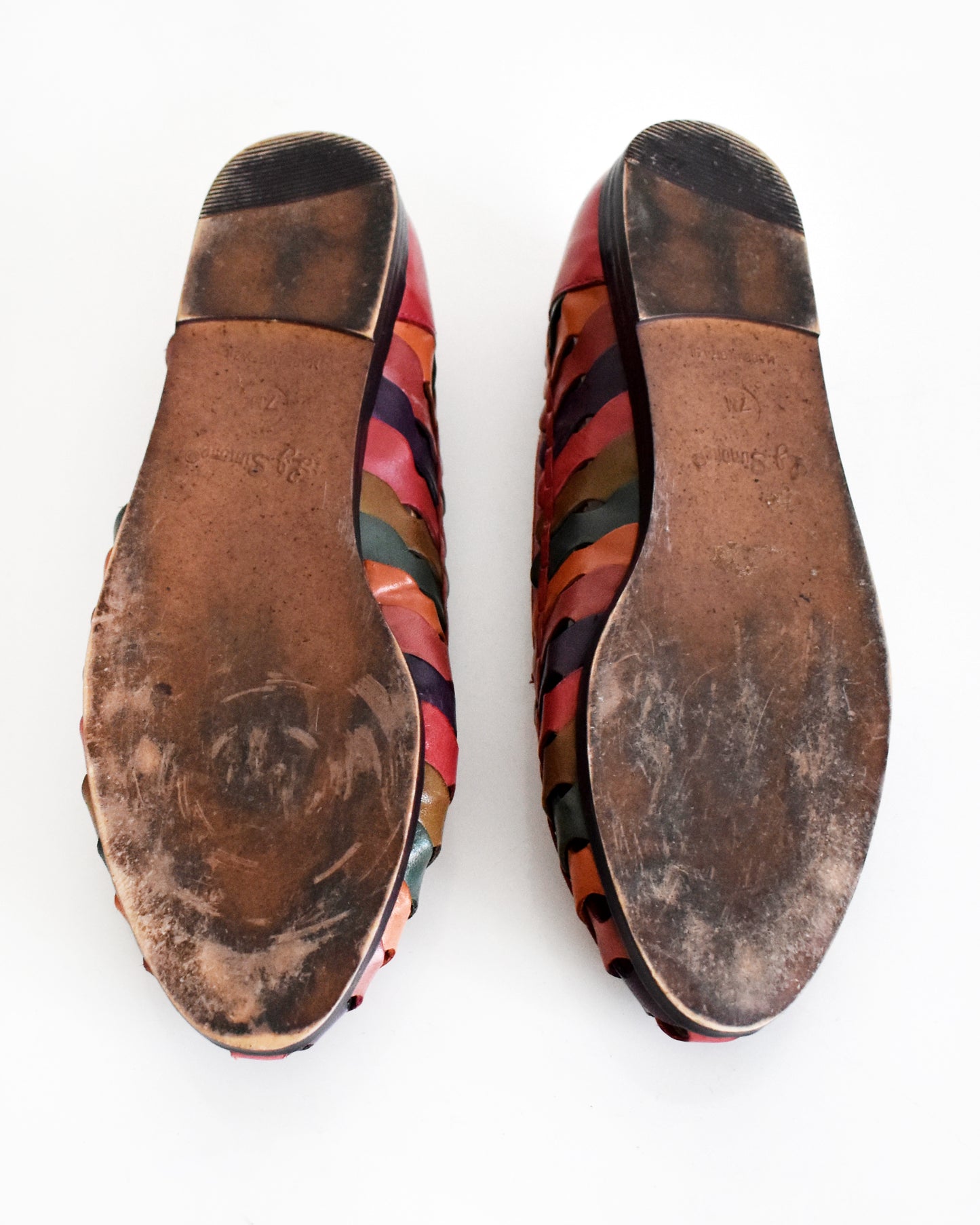 A pair of slip-on shoes shown from the bottom, featuring a brown sole and a side strap pattern in multiple colors including red, pink, orange, green, and blue, on a white background.