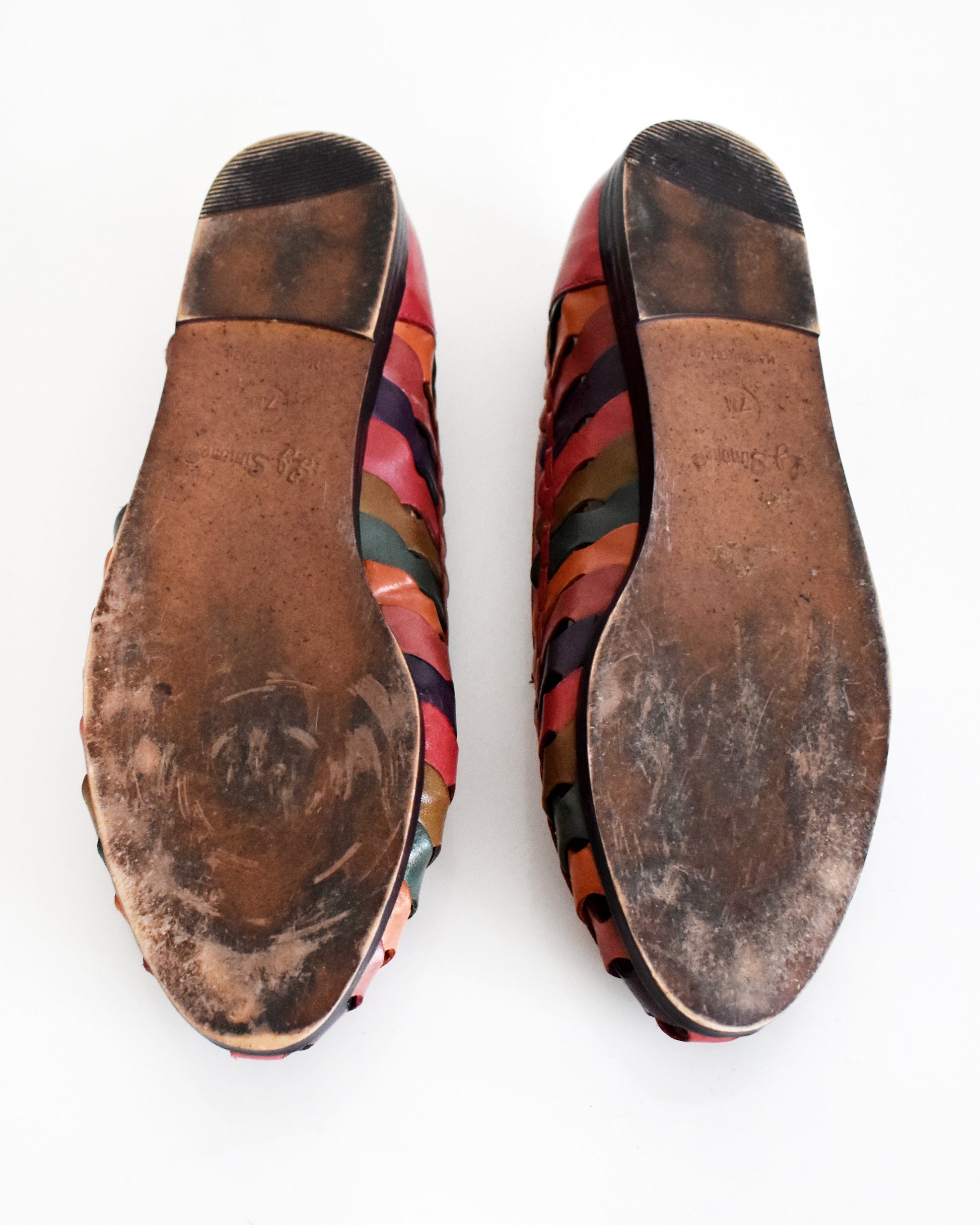 A pair of slip-on shoes shown from the bottom, featuring a brown sole and a side strap pattern in multiple colors including red, pink, orange, green, and blue, on a white background.