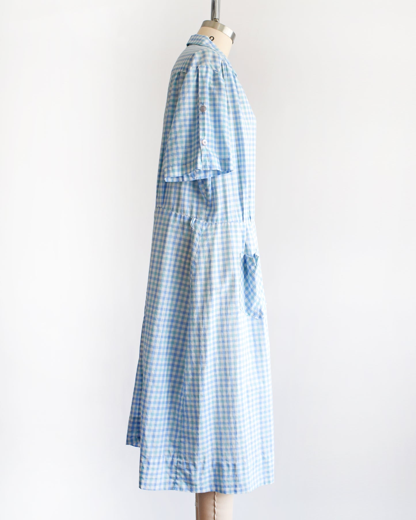 side view of a vintage 1950s light blue and white gingham short-sleeve button-front dress displayed on a mannequin against a plain white background.