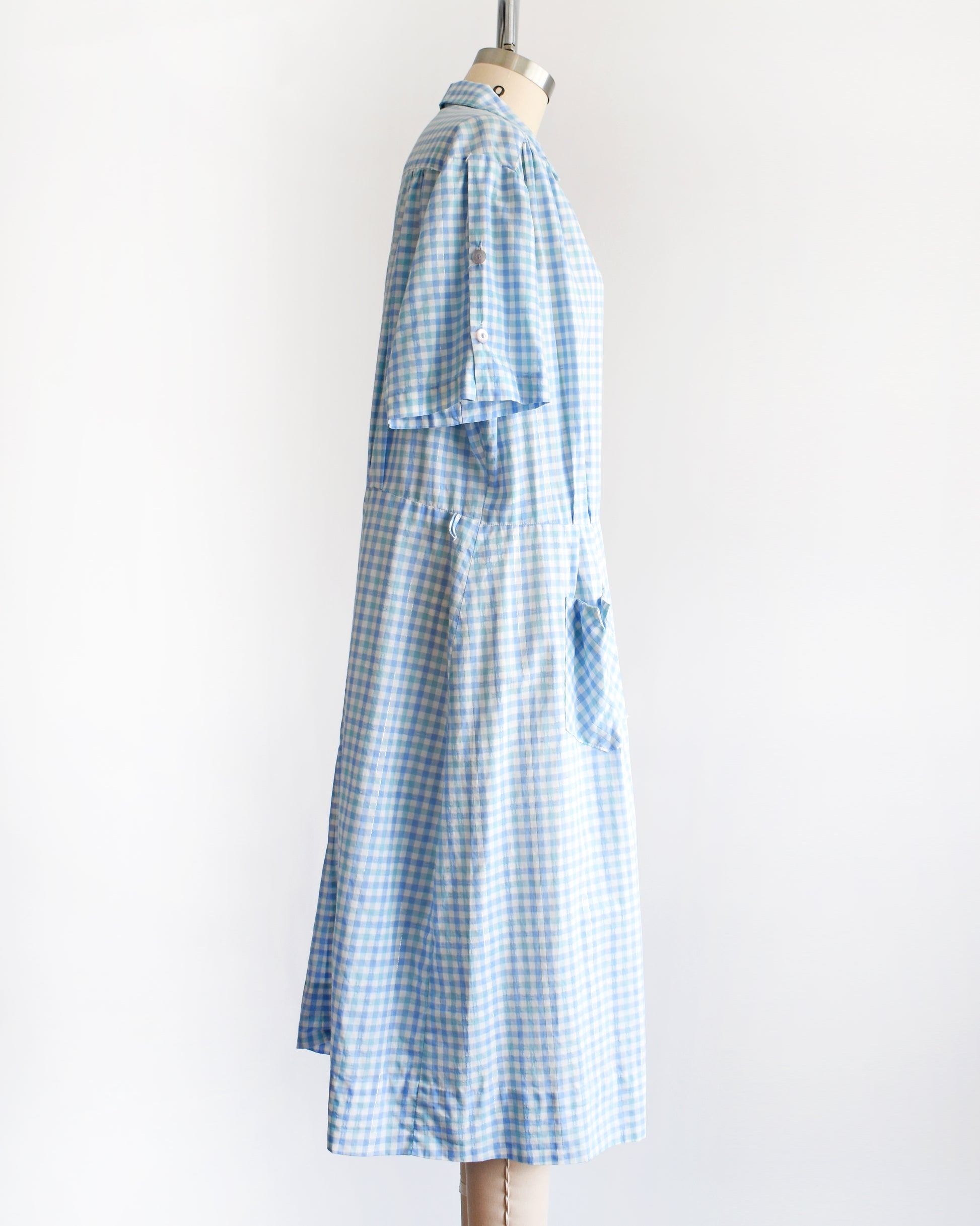 side view of a vintage 1950s light blue and white gingham short-sleeve button-front dress displayed on a mannequin against a plain white background.