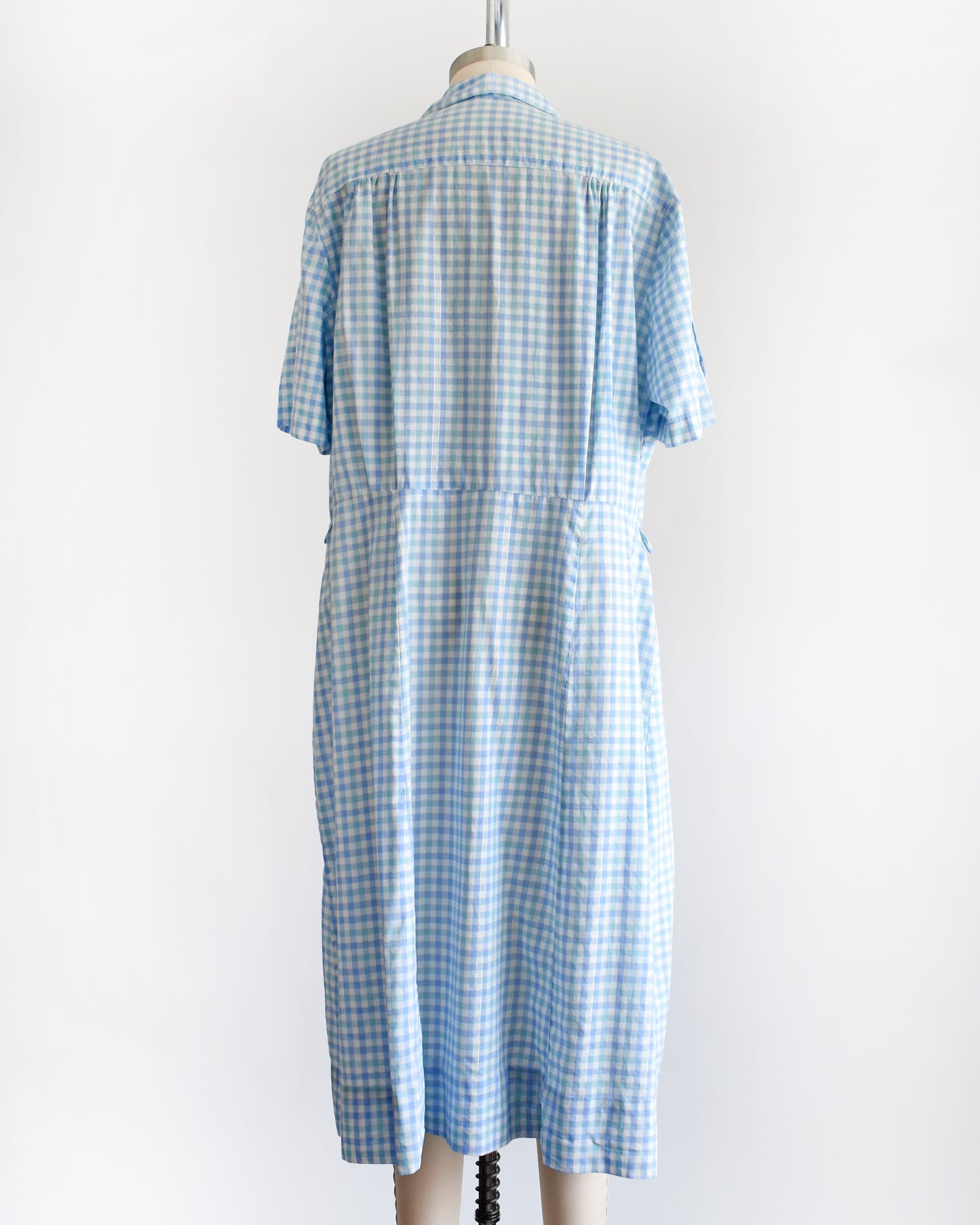back view of a vintage 1950s light blue and white gingham short-sleeve button-front dress displayed on a mannequin against a plain white background.
