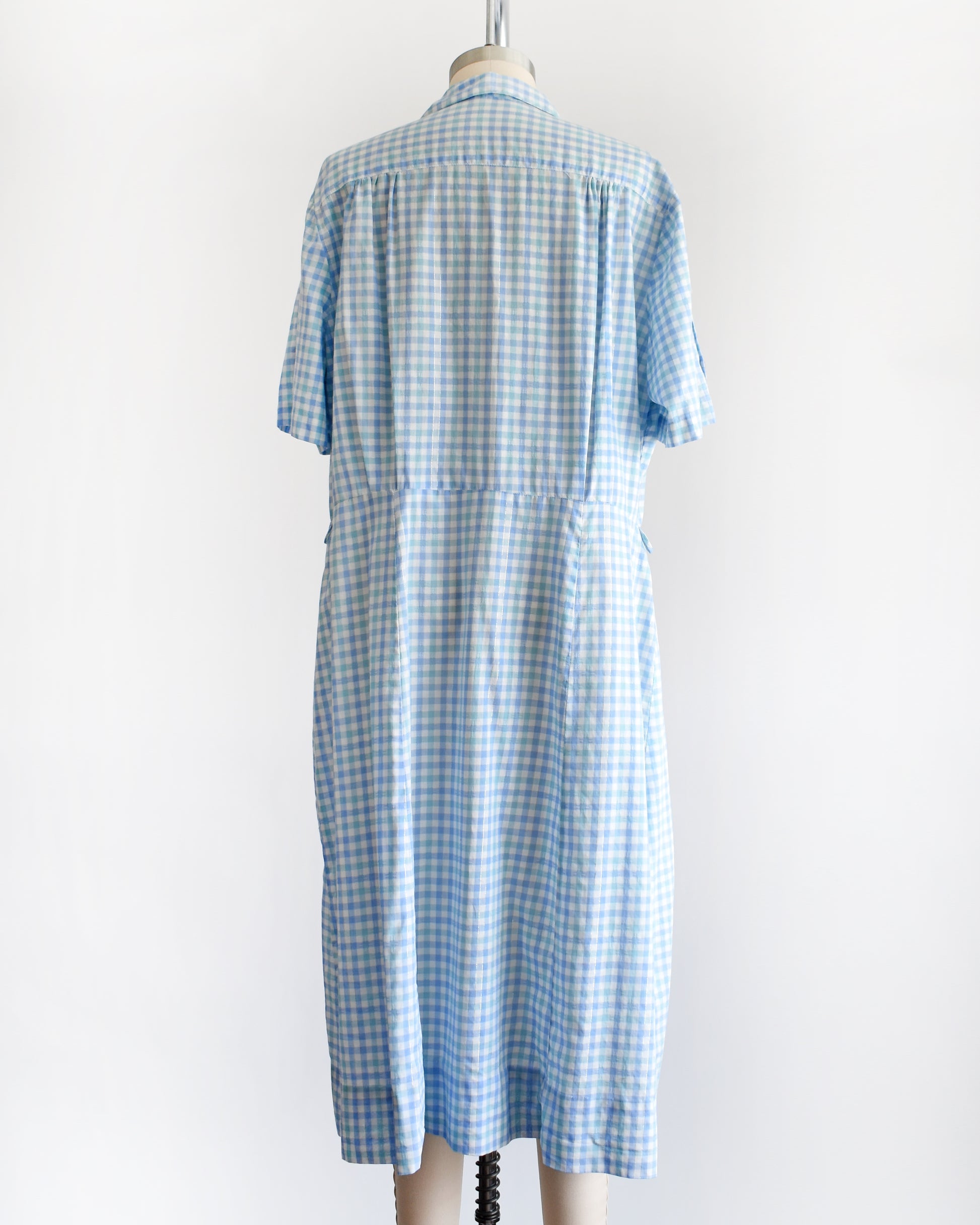 back view of a vintage 1950s light blue and white gingham short-sleeve button-front dress displayed on a mannequin against a plain white background.