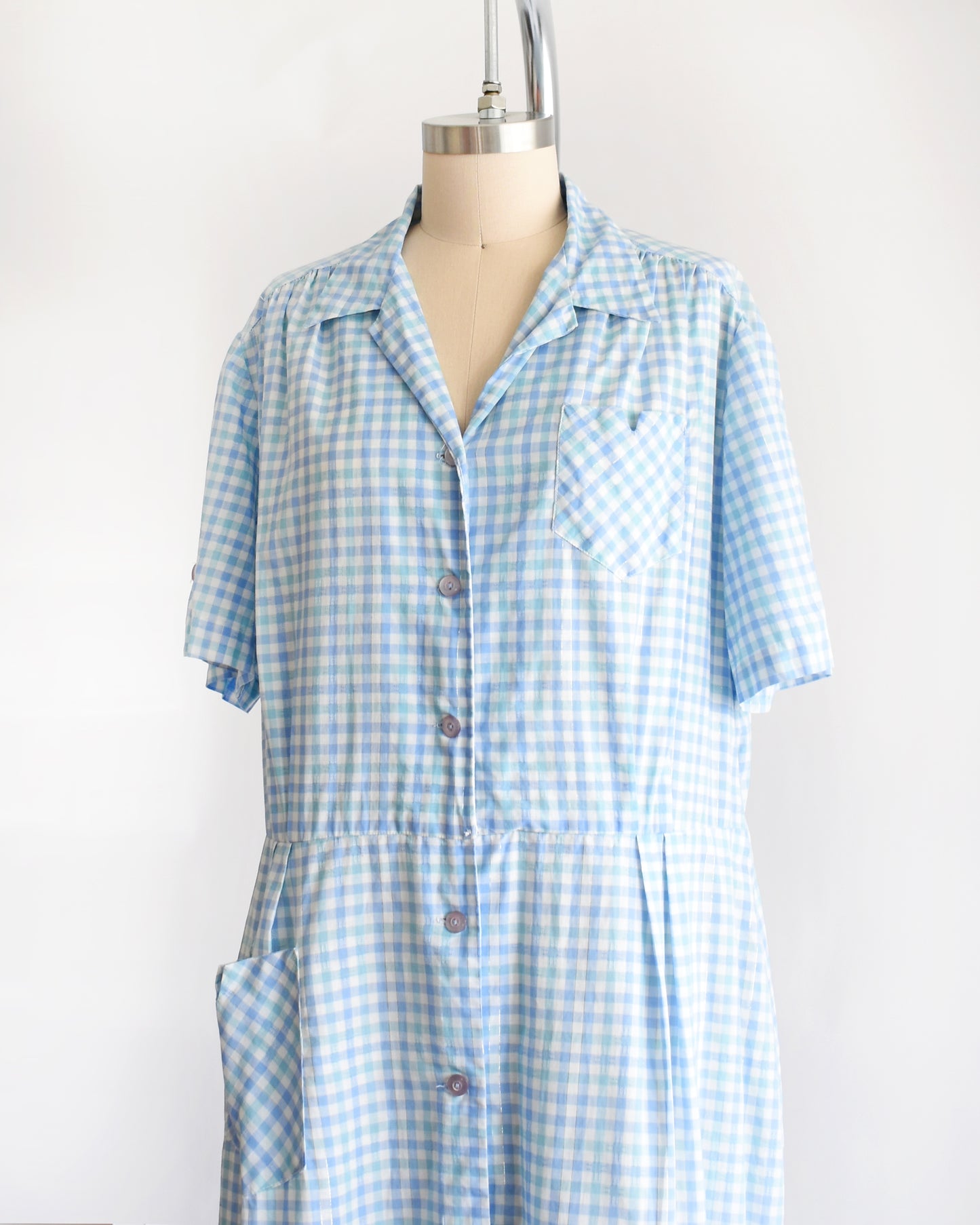 side front view of a vintage 1950s light blue and white gingham short-sleeve button-front dress displayed on a mannequin against a plain white background.