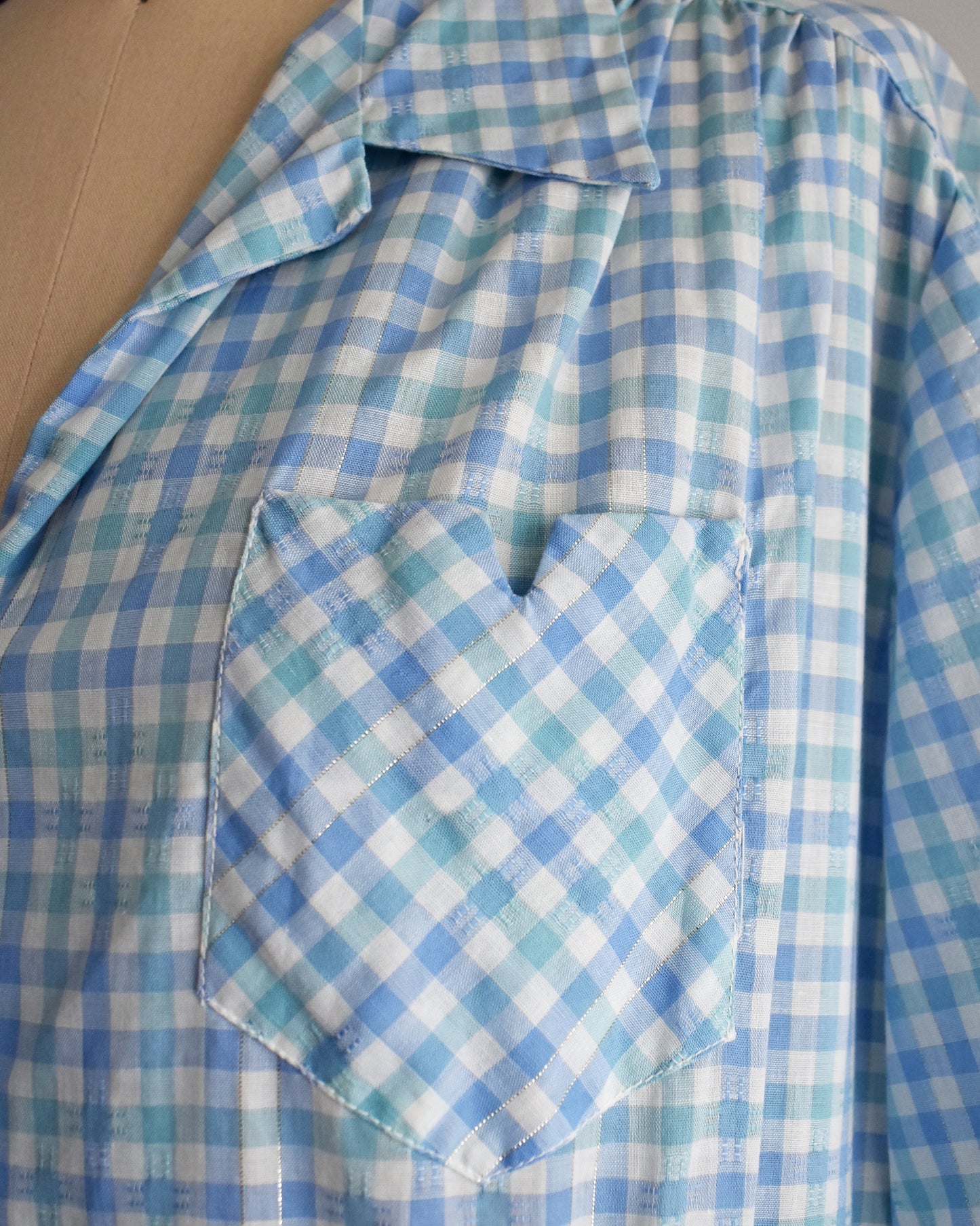 close up of the chest pocket on a vintage 1950s light blue and white gingham short-sleeve button-front dress displayed on a mannequin against a plain white background.